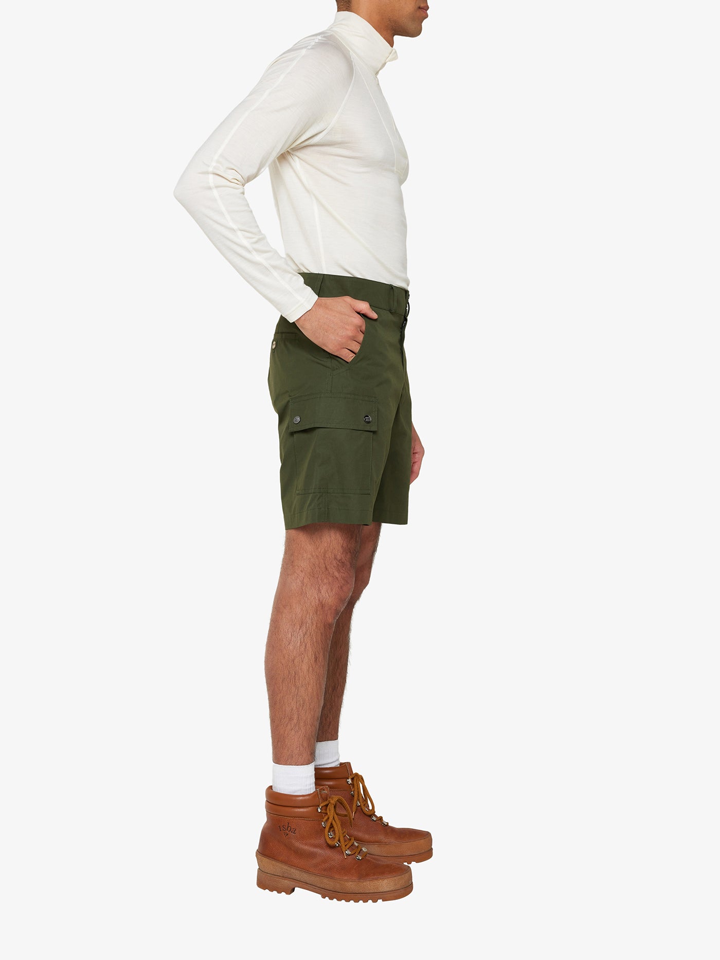 A person is standing in profile against a white background, wearing a long-sleeved, off-white Ventile® cotton fabric shirt, We Norwegians' Lyngen Shorts Men Olive Green with pocket flaps, white socks, and light brown hiking boots. The person’s left arm is bent slightly at the elbow.