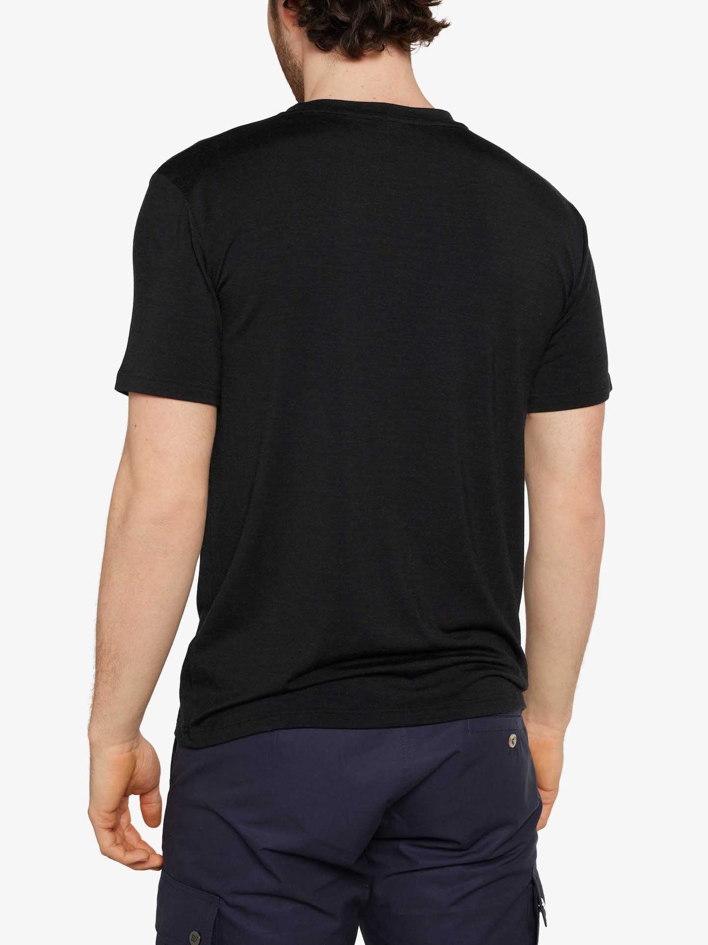 A person with short, dark hair is shown from the back, wearing a We Norwegians Hygge Tee Men Black made of super fine merino wool and navy blue pants against a plain white background. The person's arms are relaxed by their sides.