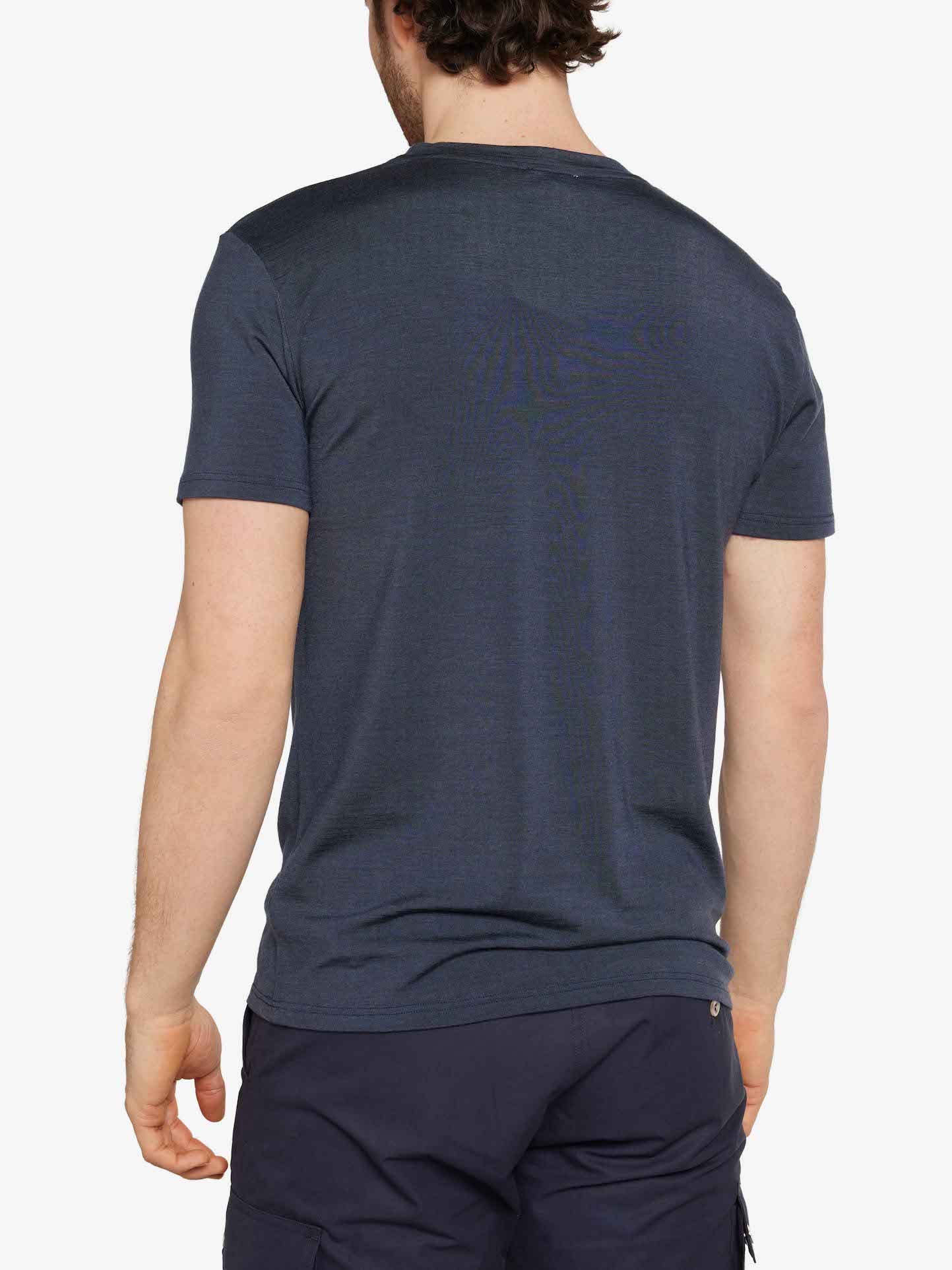 A person with short dark hair is standing with their back to the camera, wearing a dark blue Hygge Tee Men by We Norwegians, made from Merino wool, and matching dark blue shorts. The background is plain white.