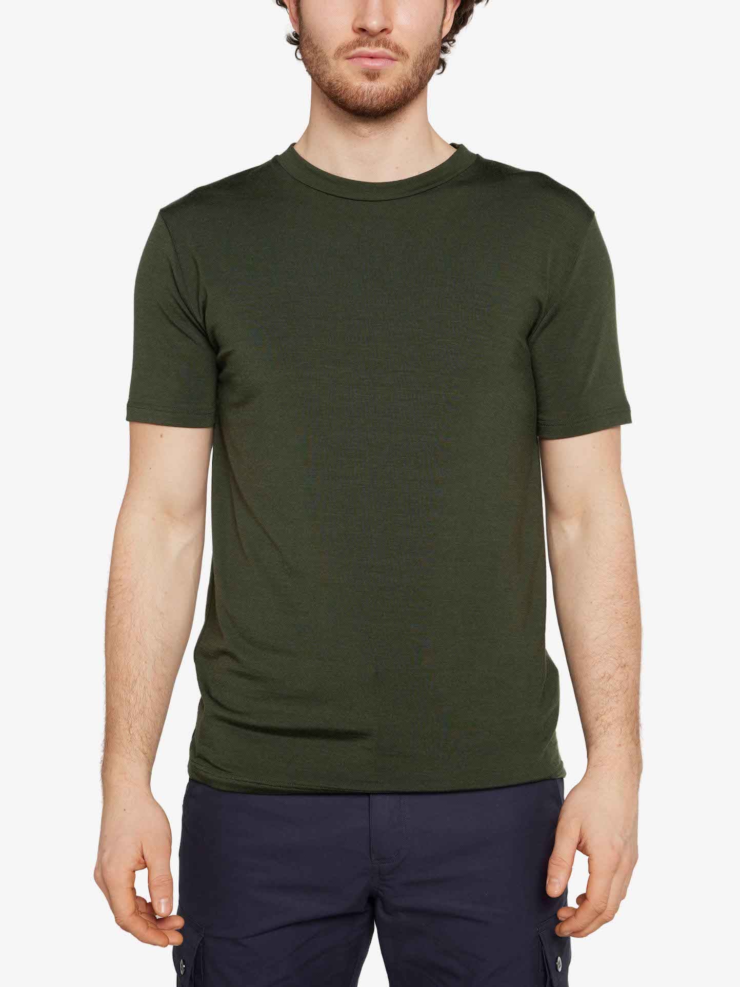 A person with short, dark hair is standing against a white background. They are wearing an olive green Hygge Tee by We Norwegians and navy blue pants. Their hands are relaxed by their sides. The person's face is not fully visible.