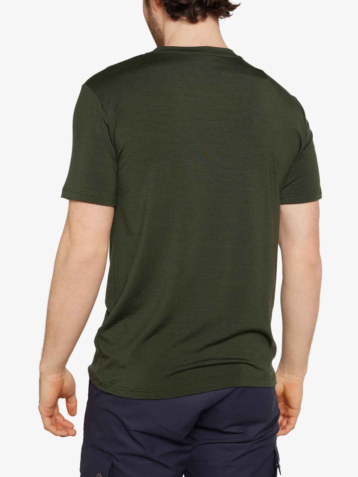 A person with short, dark hair is standing with their back to the camera. They are wearing a We Norwegians Hygge Tee Men in Olive Green and dark blue pants. The background is plain white.