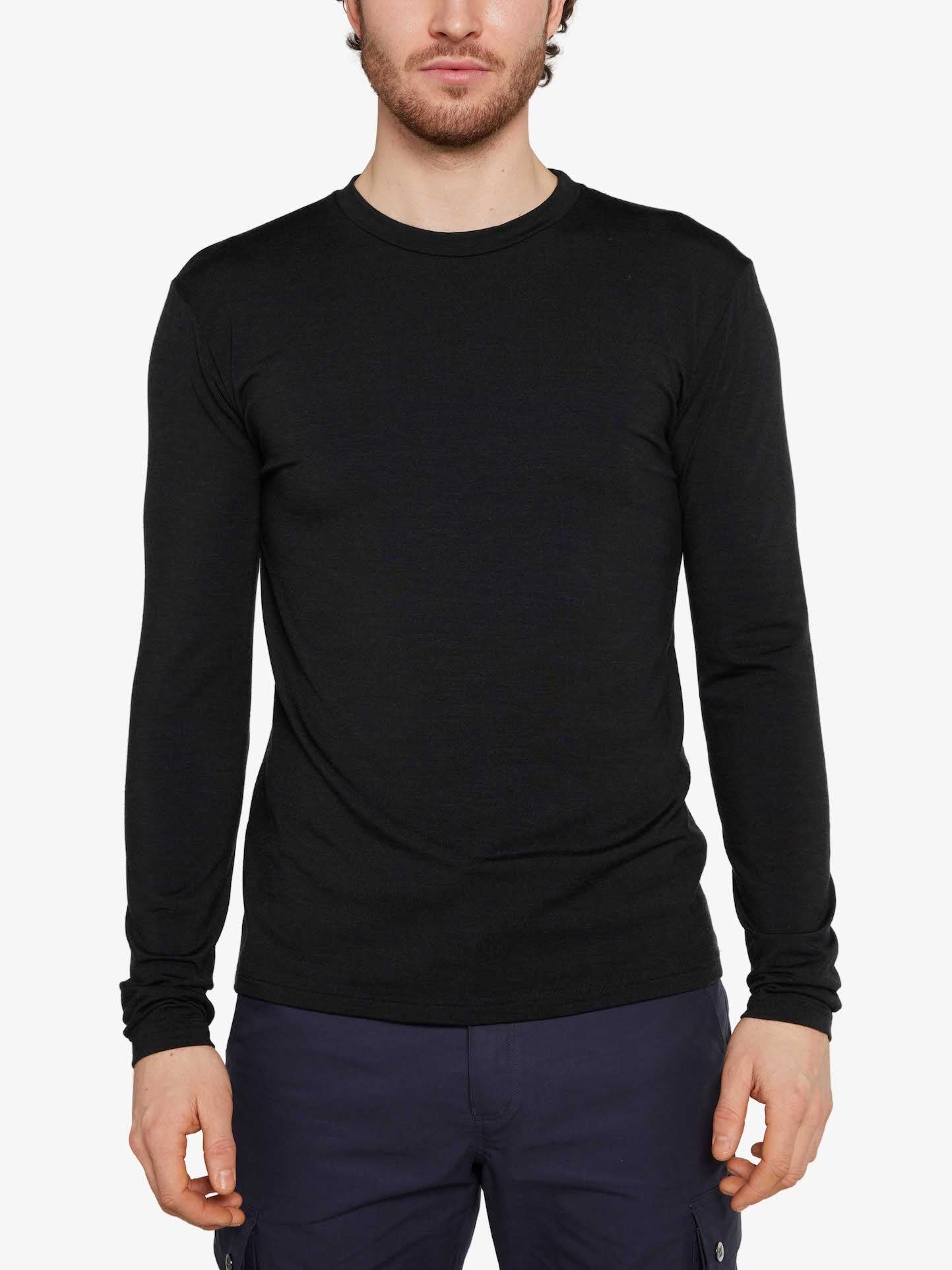 A person is wearing the Hygge LongSleeve Men by We Norwegians, a fitted long-sleeve black shirt made of super fine merino wool, paired with dark pants against a plain light grey background. The individual's face is partially visible, with emphasis on the upper body and attire.