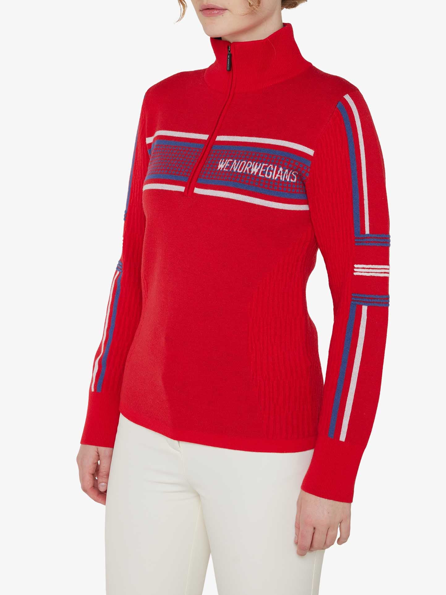 A person is wearing the Stryn Zipup Women sweater by We Norwegians. This red long-sleeve sweater features blue and white stripes on the sleeves and chest, along with the text "We Norwegians" on the front. Boasting a retro design, this classic ski sweater includes a high collar with a partial front zipper. The person is also wearing white pants.