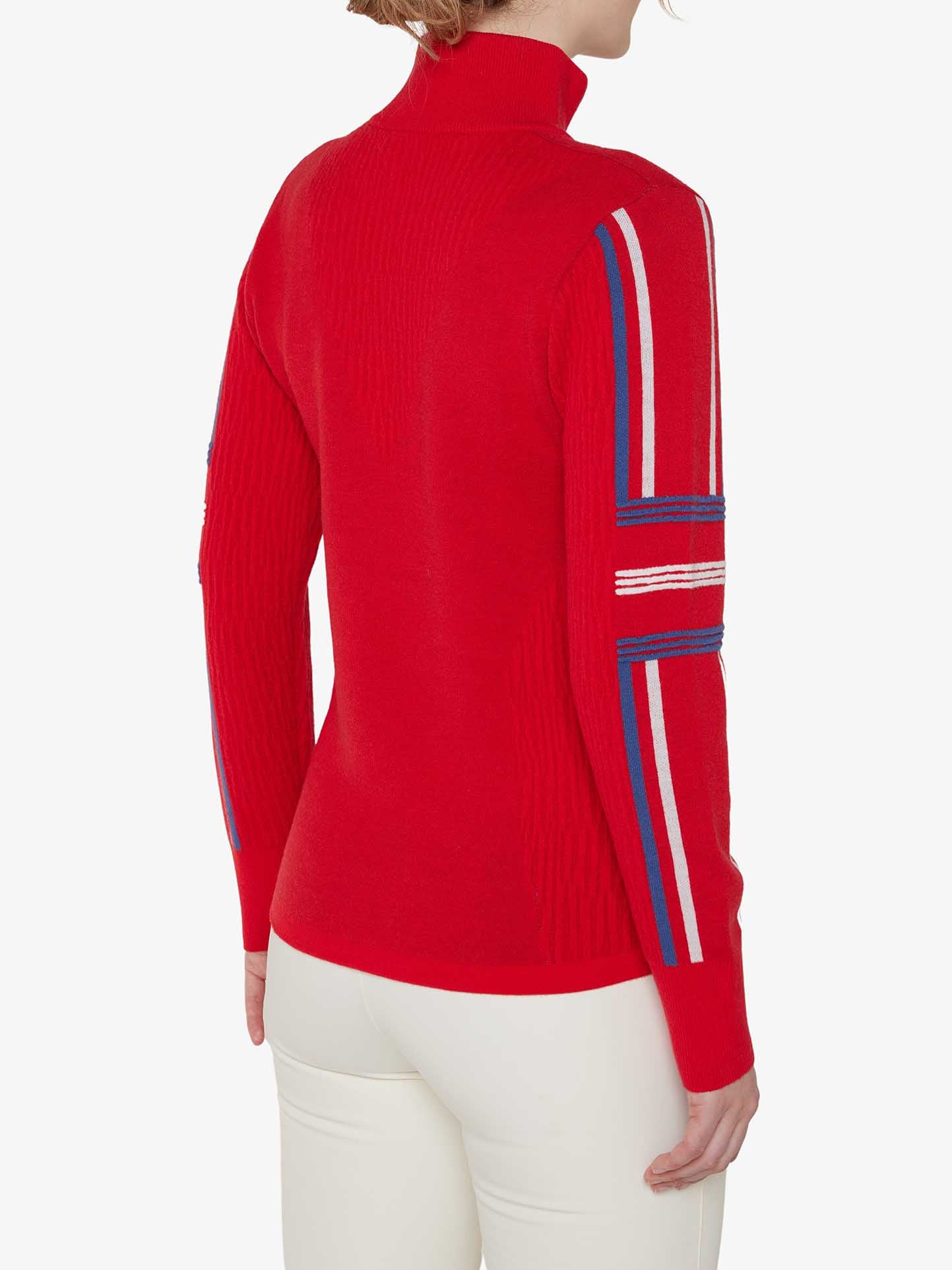 A person wearing the We Norwegians Stryn Zipup Women sweater, which features a red long-sleeved design with blue, white, and red striped detailing on the sleeves. The sweater has a classic ski style with a turtleneck and is paired with white pants. The person is shown from the back.