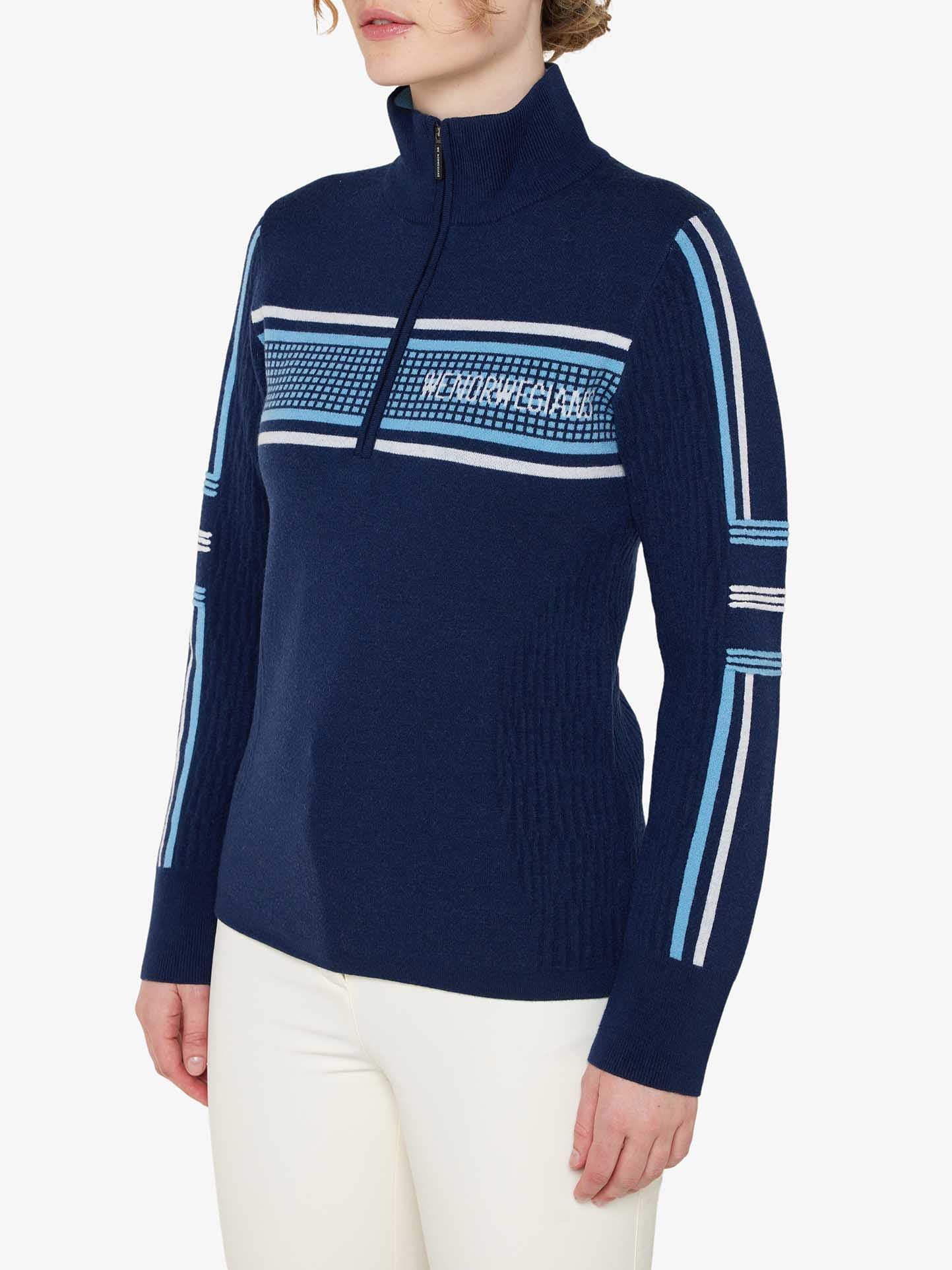 A person wearing the We Norwegians Stryn Zipup Women, a navy blue long-sleeve classic ski sweater with light blue and white horizontal stripes across the chest, arms, and shoulders. The retro design features a half-zip front and a high collar. They are also wearing white pants against a plain white background.