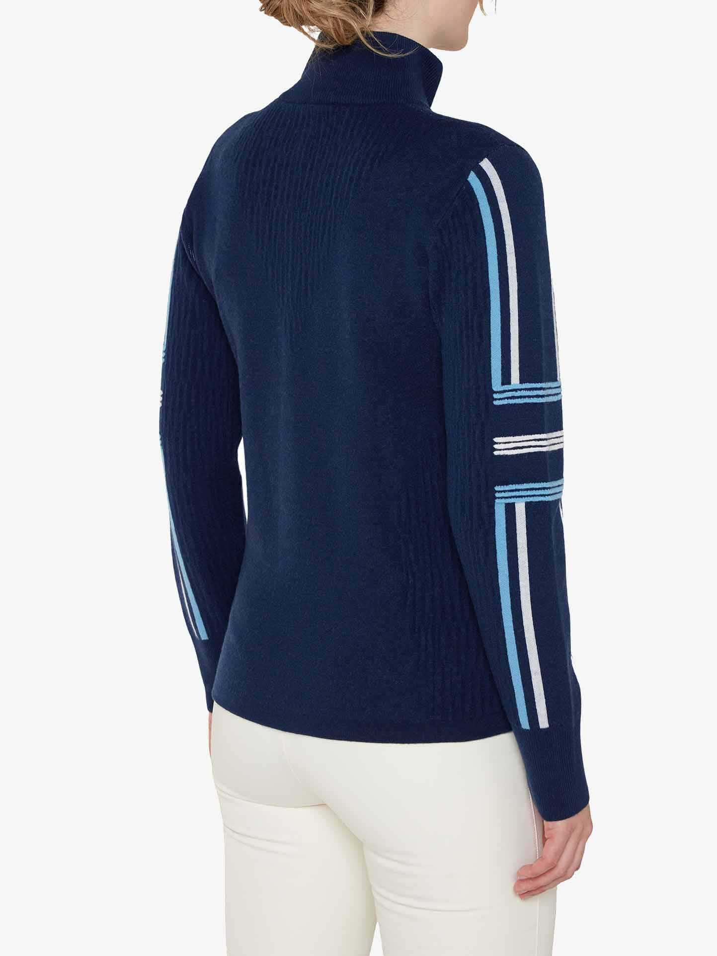 A person stands facing away from the camera, wearing a dark blue Stryn Zipup Women turtleneck by We Norwegians, featuring light blue and white stripes on the sleeves that evoke the style of a classic ski sweater. They are also sporting white pants. The background is plain white.