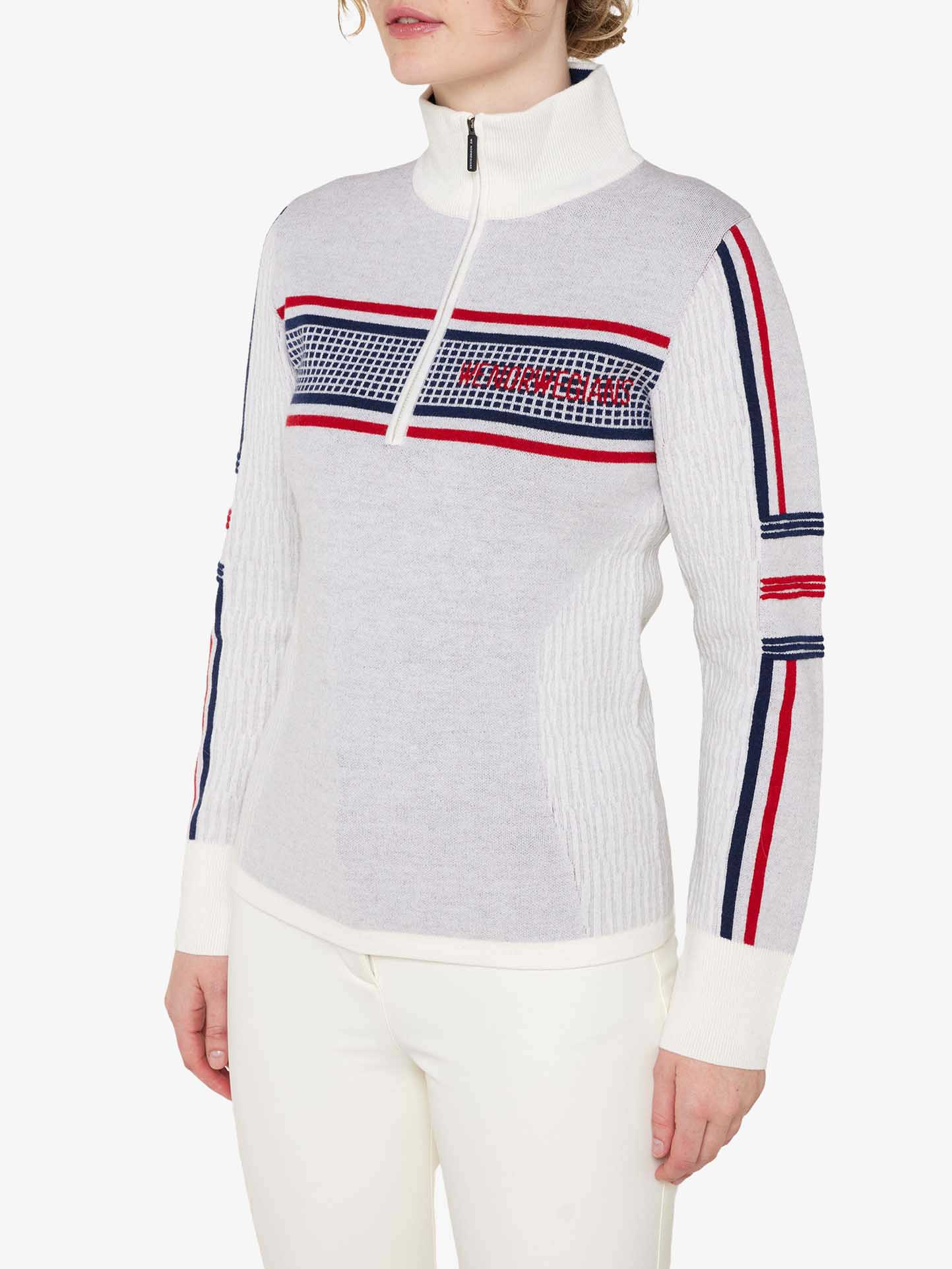 A person is wearing the Stryn Zipup Women sweater by We Norwegians, which features a light gray color with long sleeves and a high collar. The classic ski sweater has a front half-zip closure and showcases red, white, and blue horizontal stripes on the chest and sleeves, along with a small grid pattern on the chest. The person is also wearing white pants.