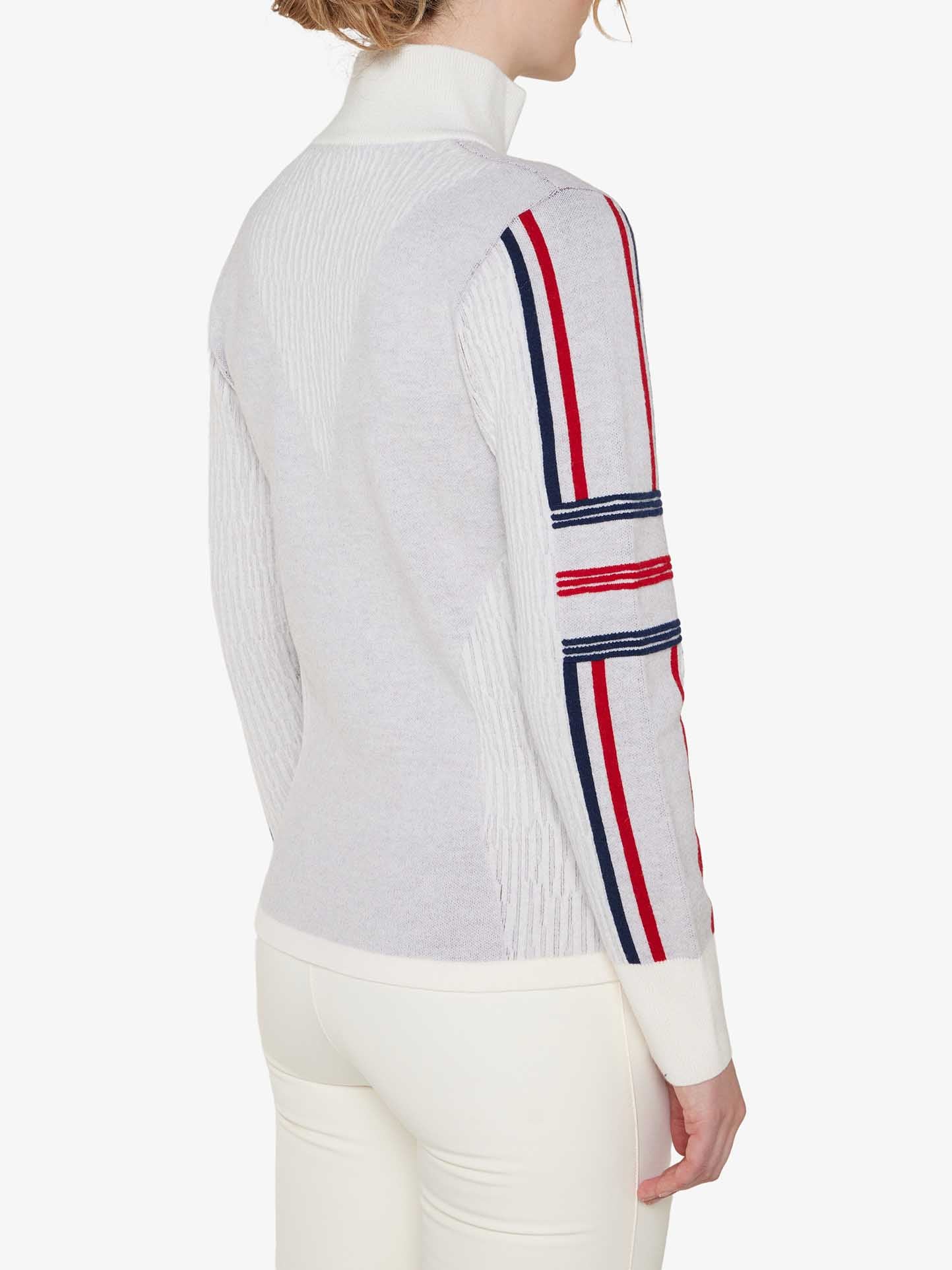 A person is wearing the Stryn Zipup Women by We Norwegians, a light gray, long-sleeved classic ski sweater with red and blue striped detailing on the sleeves. Made from ultrafine merino wool, the sweater features a high collar. The person is turned slightly away from the camera, showcasing the back and side profiles of the attire.