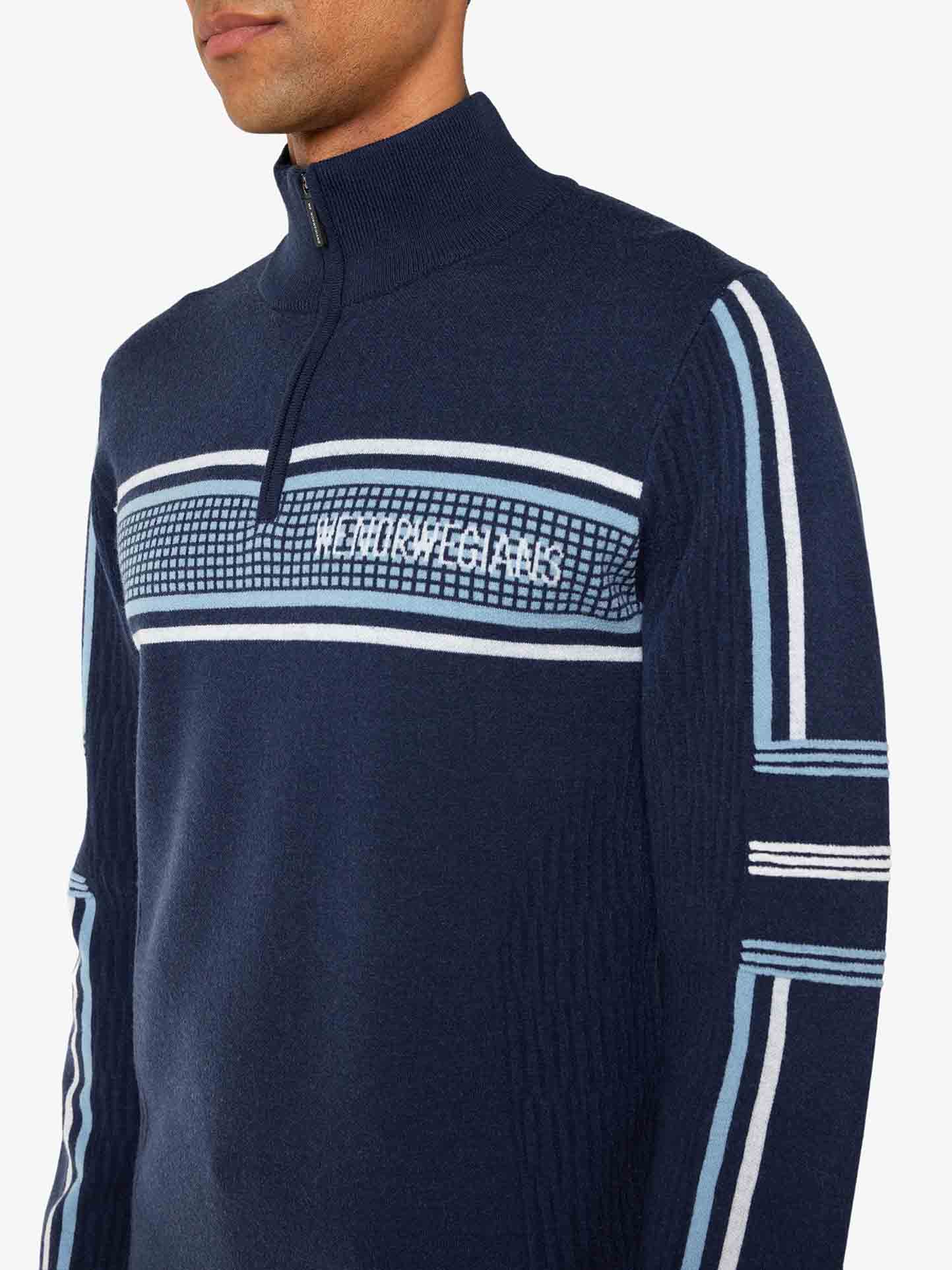 A person is wearing a Stryn Zipup Men sweater by We Norwegians, featuring navy blue ultrafine merino wool with light blue and white stripes on the sleeves and chest. It has a quarter-zip turtleneck and a geometric pattern across the chest, with text that appears to read "NEO FUTURISTIC.