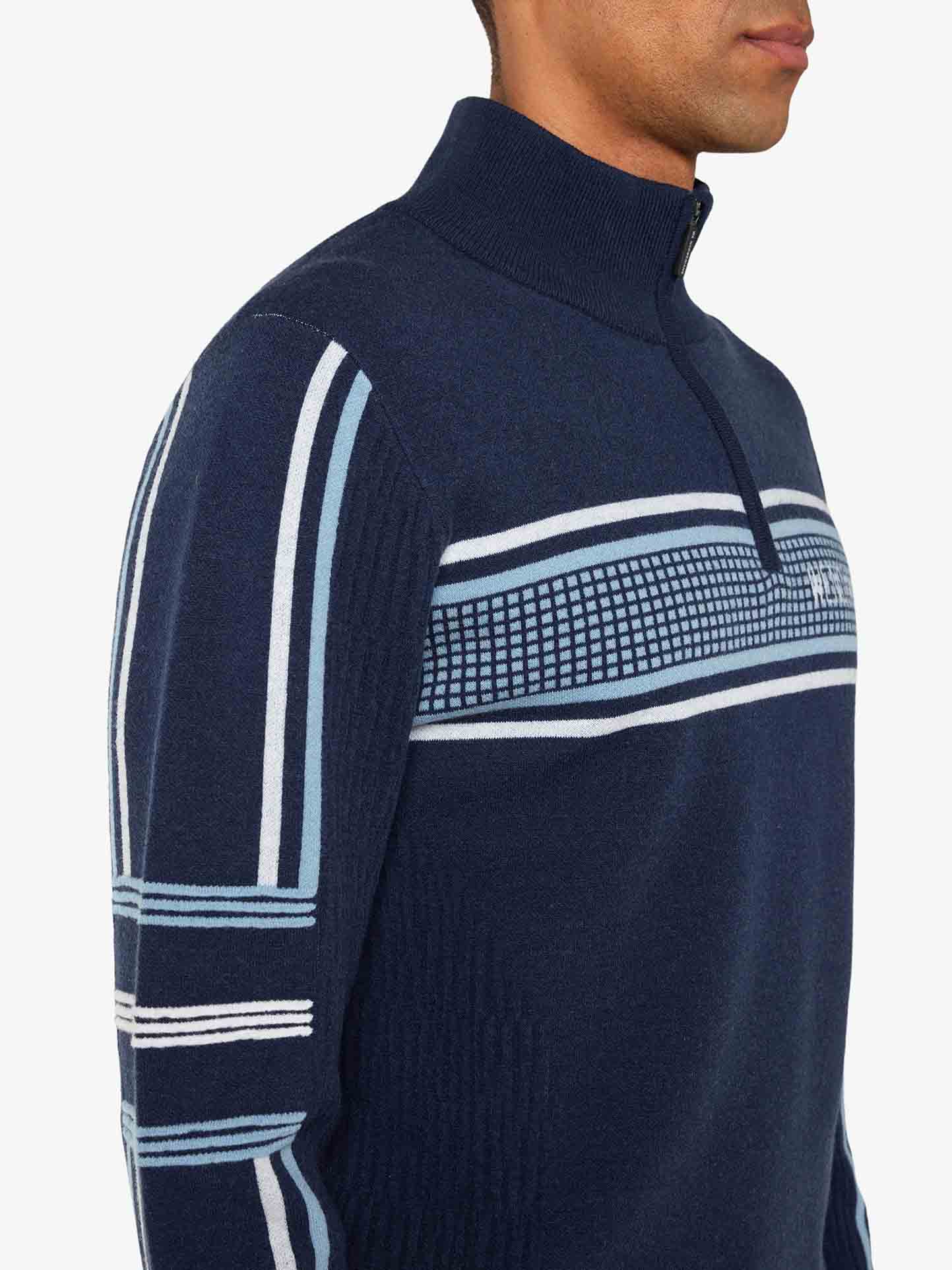 A person is wearing the Stryn Zipup Men, a navy blue half-zip retro ski sweater by We Norwegians, featuring light blue and white geometric patterns across the chest and sleeves. The sweater has a ribbed stand collar and boasts a textured knit design in ultrafine merino wool. In the close-up side profile view, the person's face is not visible.