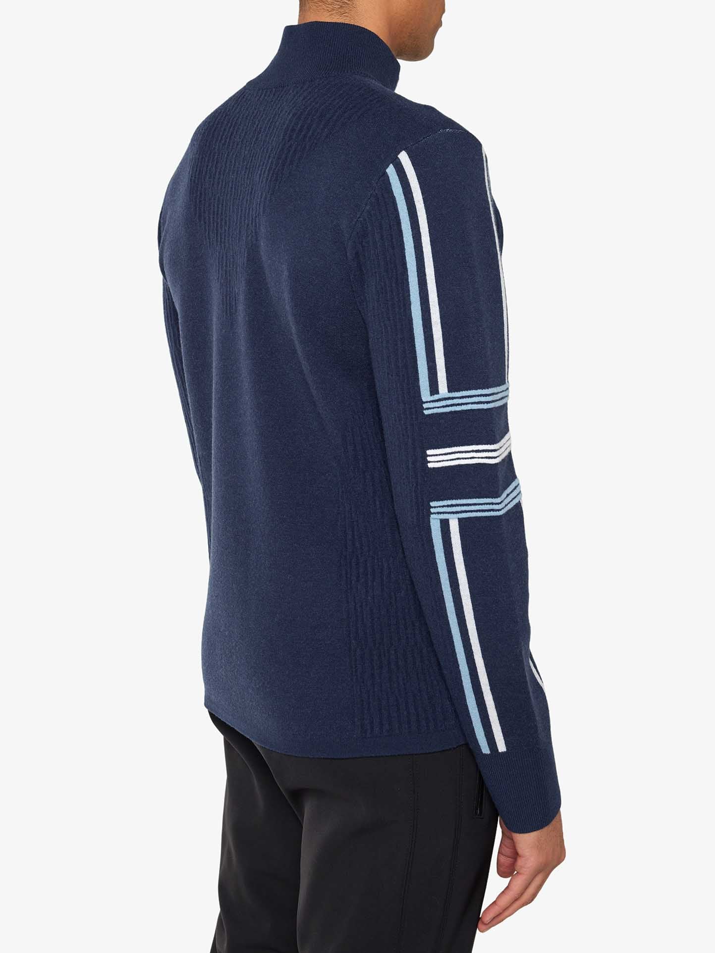 A person is wearing the Stryn Zipup Men by We Norwegians, a navy blue retro ski sweater with light blue and white stripes on the sleeves. The high-collared sweater is seen from the back and side, paired with black pants.