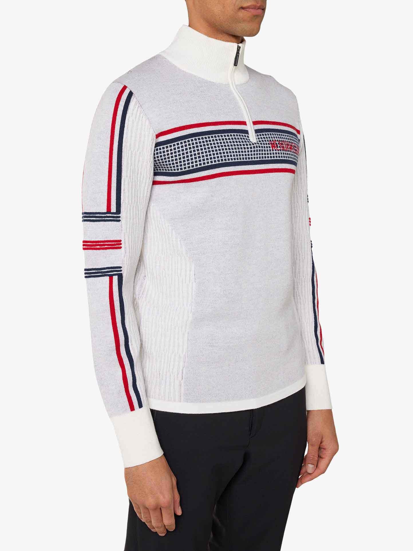 A person is wearing a We Norwegians Stryn Zipup Men sweater, featuring red and blue horizontal stripes on the chest and sleeves. This long-sleeved ultrafine merino wool garment boasts a half-zip collar. The individual pairs the sweater with dark pants against a plain white background.