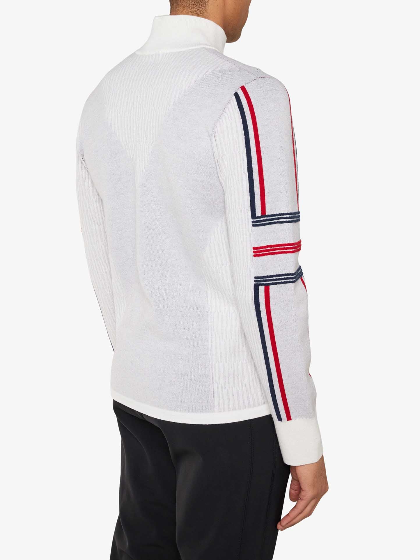 A man, seen from behind, is wearing the Stryn Zipup Men by We Norwegians— a retro ski sweater crafted from ultrafine merino wool. The sweater features red, white, and blue striped cuffs on the left arm and a white collared neckline. He is also dressed in black pants against a plain white background.