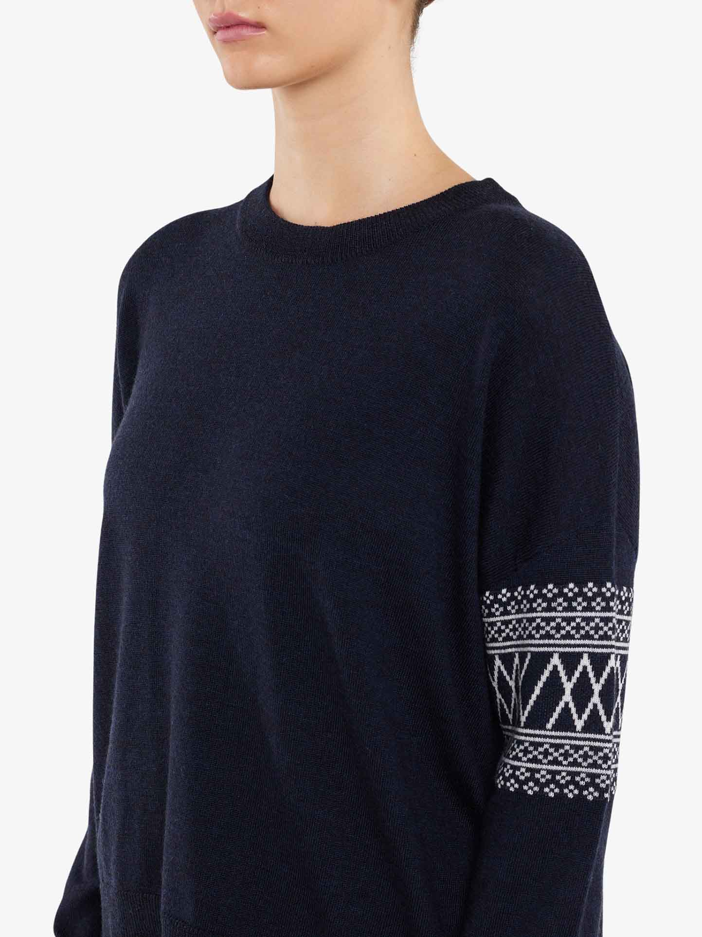 A person wearing the We Norwegians Signature Sweater Women, an over-sized dark navy blue sweater featuring a white geometric Setesdal pattern on the left sleeve. The background is plain white, and only the upper half of the person is visible, with their head turned slightly to the side.