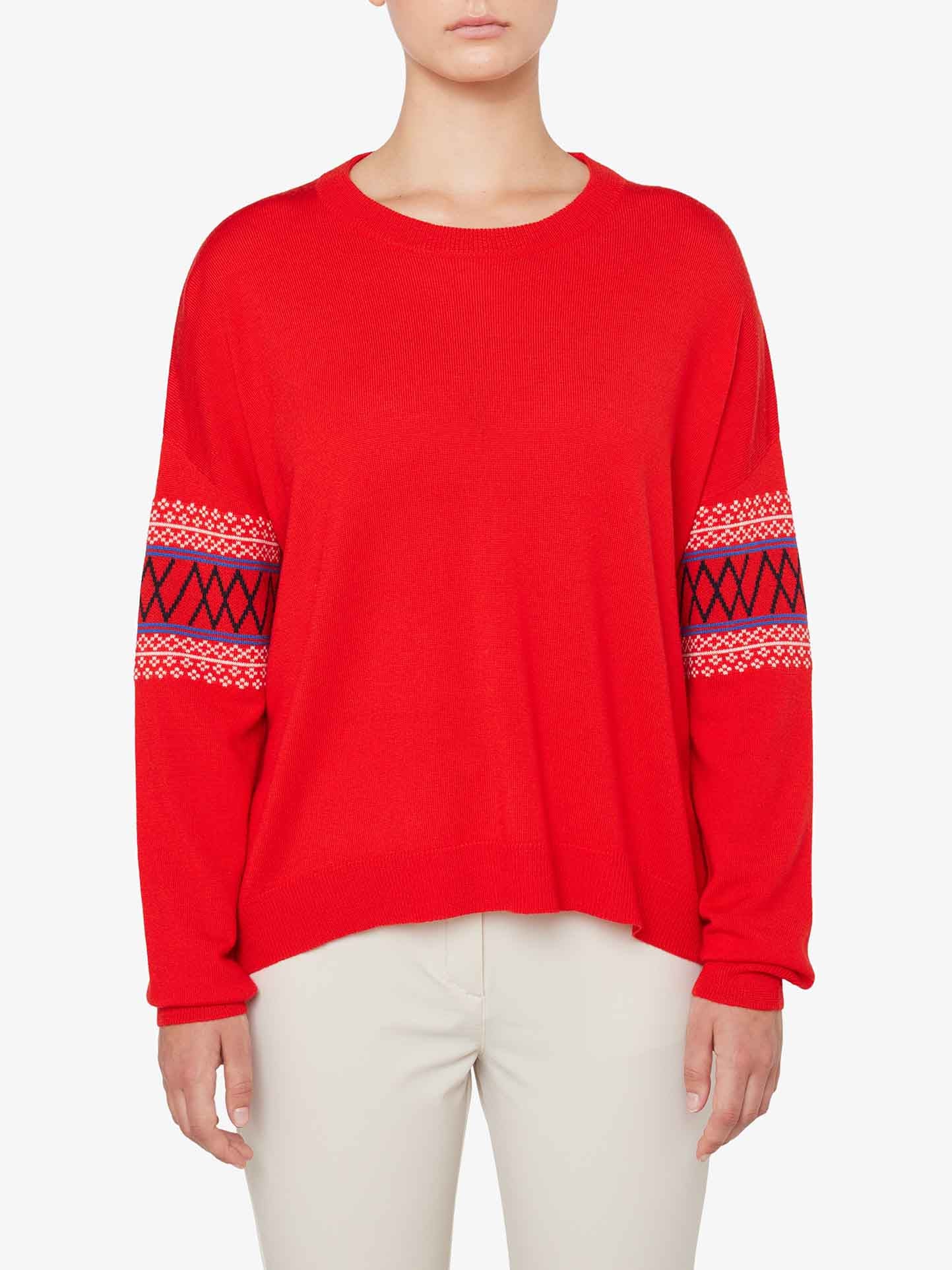 A person is wearing the We Norwegians Signature Sweater Women, an over-sized red sweater featuring intricate black, red, and white Setesdal patterns on the sleeves. They are also dressed in light-colored pants and standing against a plain white background. The Merino wool sweater features a simple round neckline and long sleeves.