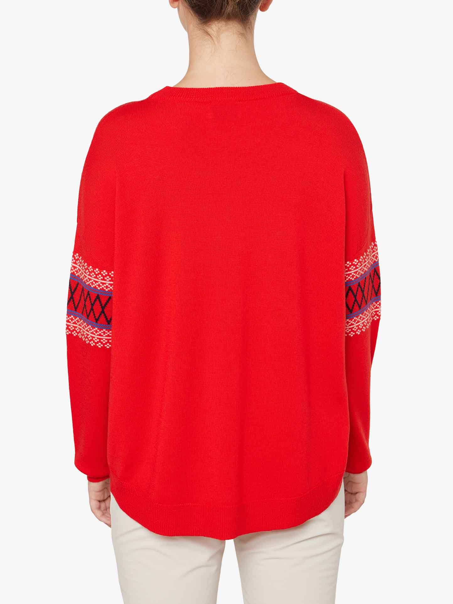 A person is shown from the back wearing an over-sized, bright red Signature Sweater Women by We Norwegians, featuring Setesdal pattern details in black and white on the sleeves. The person has light-colored pants, and the background is plain white.