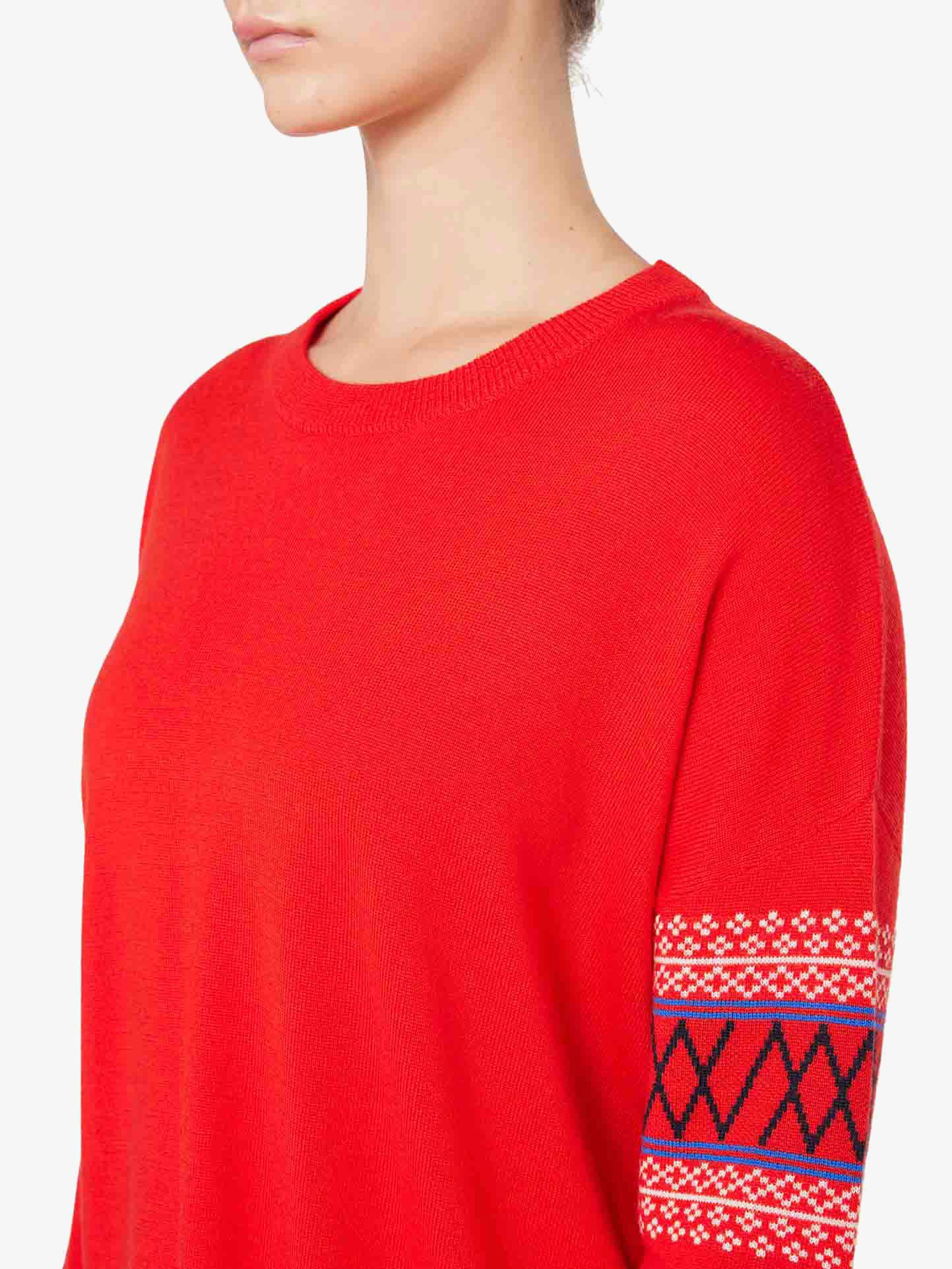 A person wearing the Signature Sweater Women by We Norwegians, an over-sized bright red Merino wool sweater with geometric detailing on the upper sleeve. The sweater features a round neckline and includes a pattern with geometric shapes in blue, white, and black. The person is shown from the shoulders up, facing sideways.