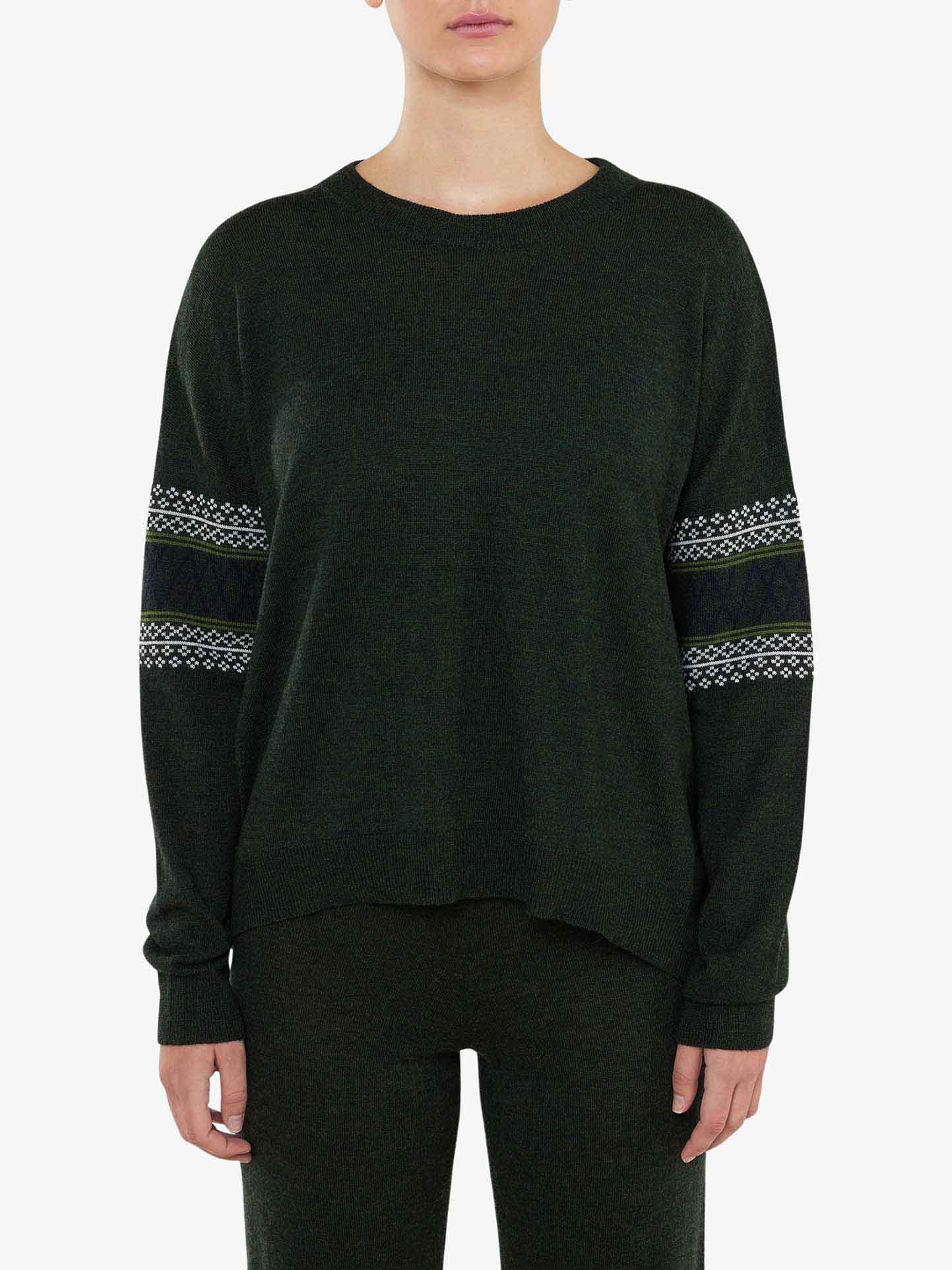 A person wearing an over-sized Signature Sweater Women by We Norwegians. The dark green, long-sleeve sweater features Setesdal pattern bands around the upper arms and a crew neckline, paired elegantly with matching dark green pants. The background is a plain light color.