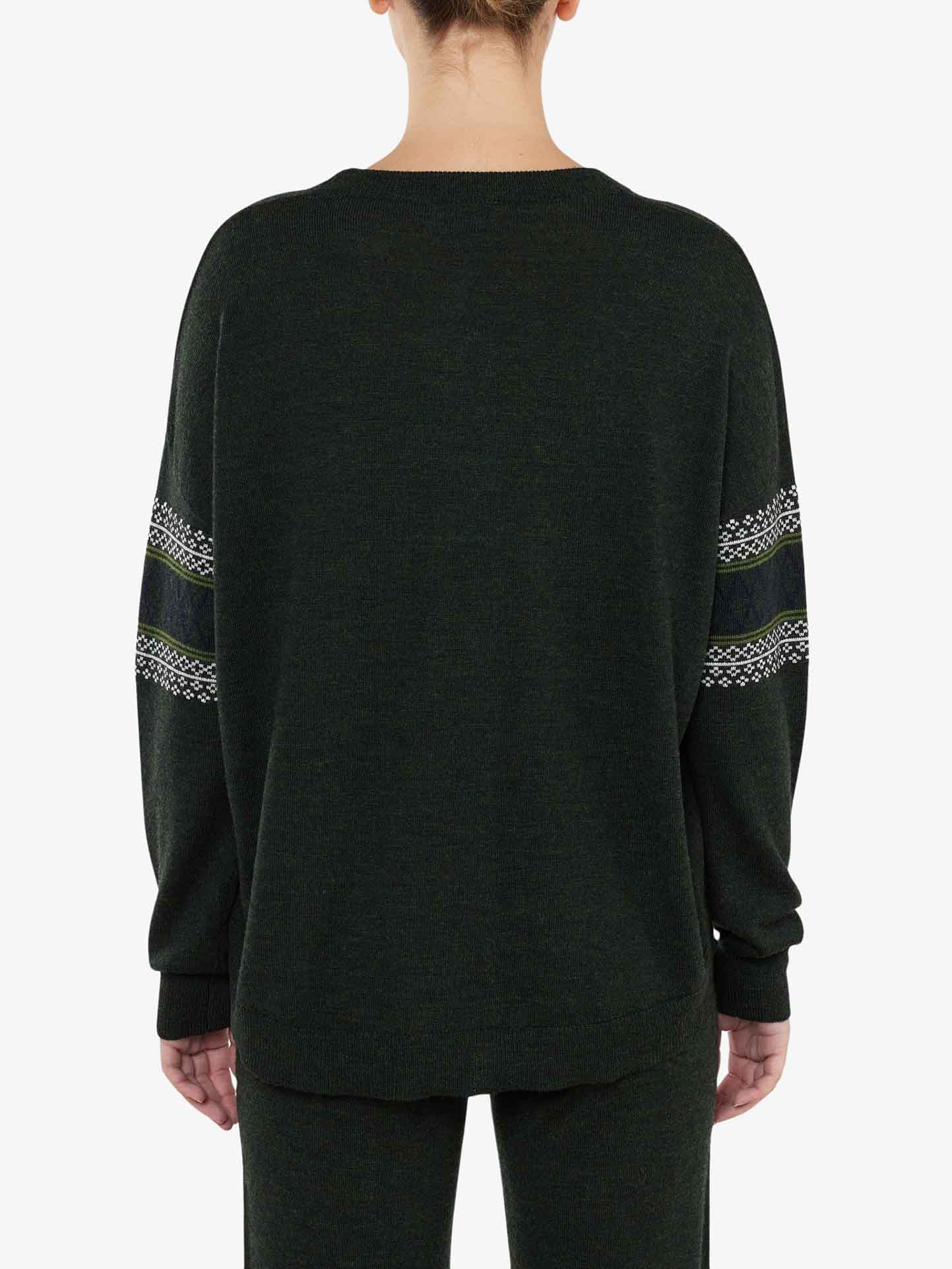 A person wearing a dark green, over-sized Signature Sweater Women by We Norwegians with decorative Setesdal patterns on the sleeves stands with their back to the camera. The Merino wool sweater has a loose fit and the person's hair is tied up. The background is plain white.