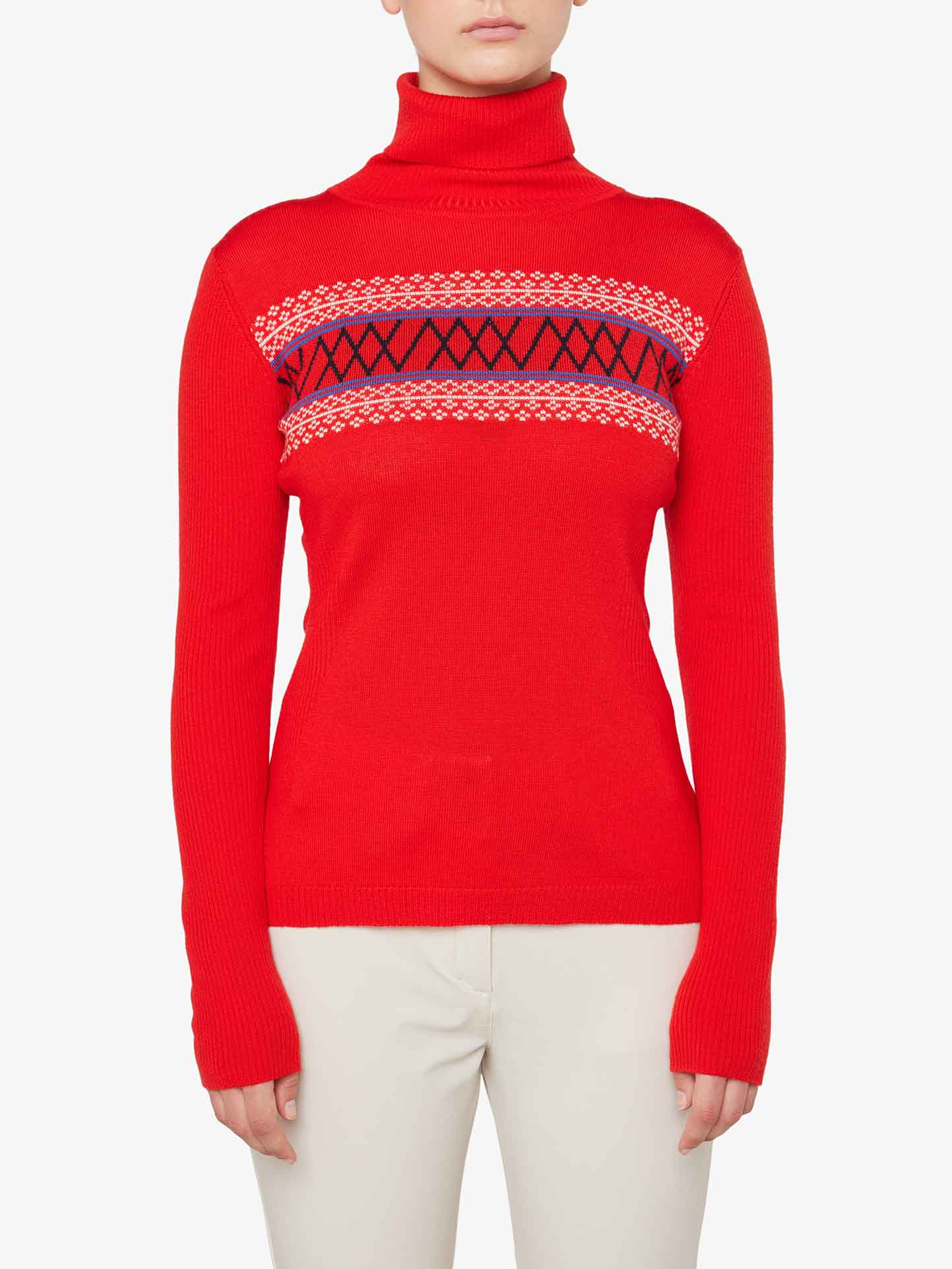 A person wearing the We Norwegians Signature Rollneck Women, a bright red top featuring a black and white patterned band across the chest, paired with light-colored pants. The sweater, with its fitted design and long sleeves, evokes retro ski outfit vibes against a plain white background.