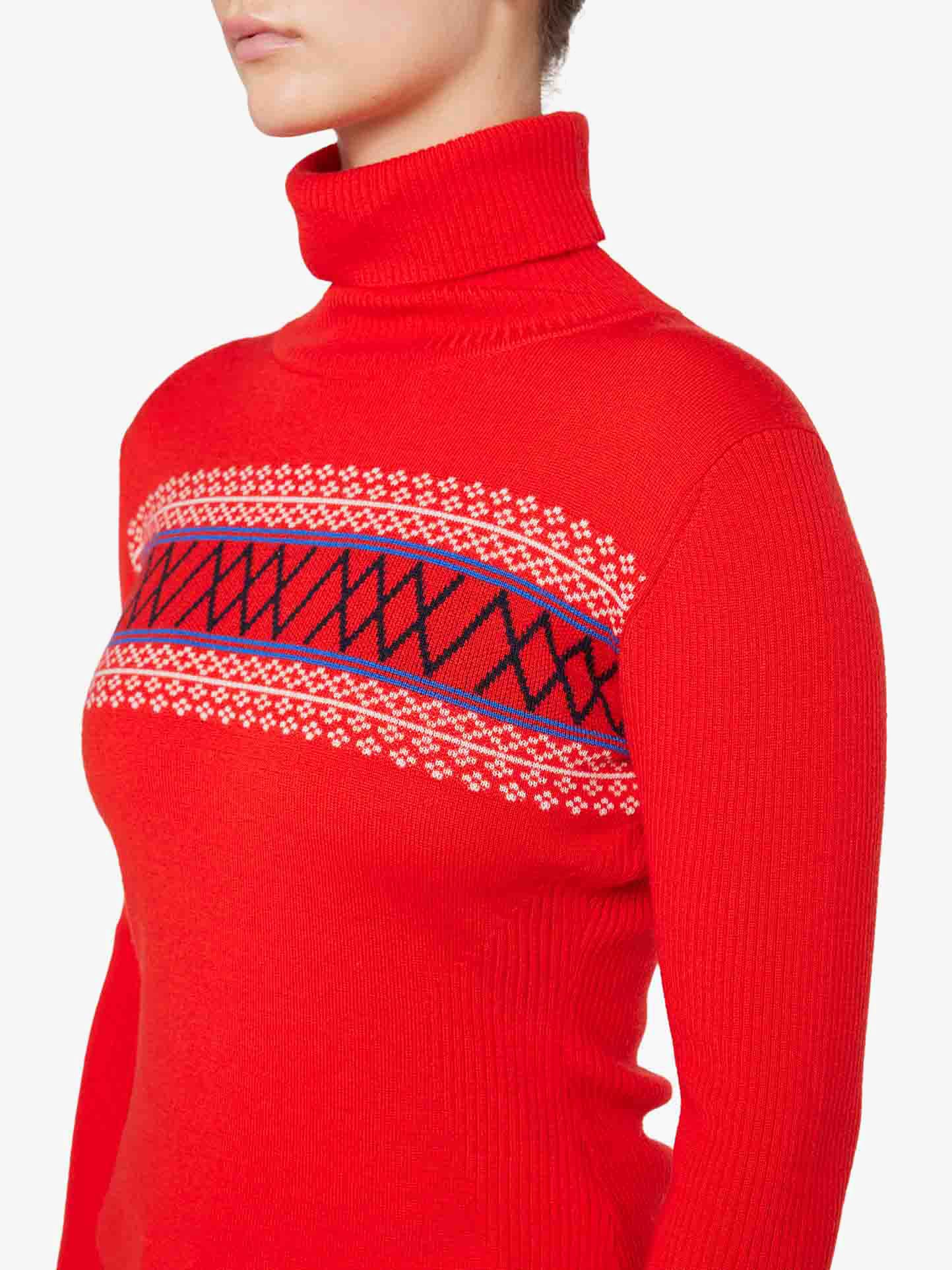 A person is wearing the "Signature Rollneck Women" top by We Norwegians, featuring a vibrant red color and adorned with a geometric pattern across the chest. The intricate design includes accents of white, black, and blue, reminiscent of classic retro ski attire. Only the lower part of their face is visible against a plain white background.