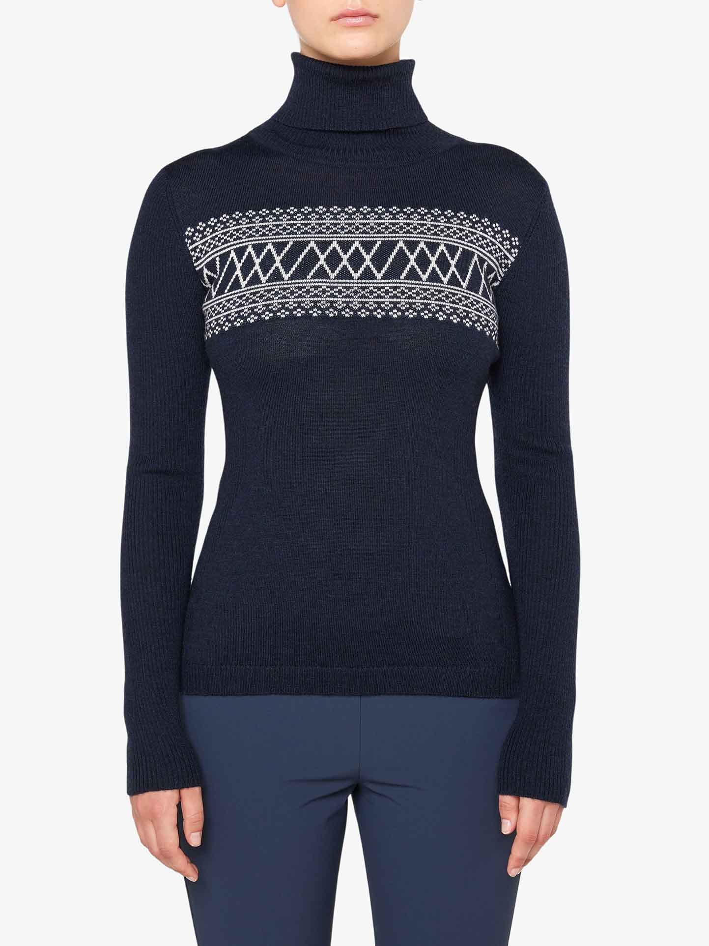 A person is wearing the "Signature Rollneck Women" from We Norwegians, a dark blue top adorned with white geometric patterns across the chest and upper arms. Made from fine Merino wool, it features long sleeves with thumbholes. Paired with dark blue pants, this ensemble evokes retro ski outfits against a plain white backdrop.