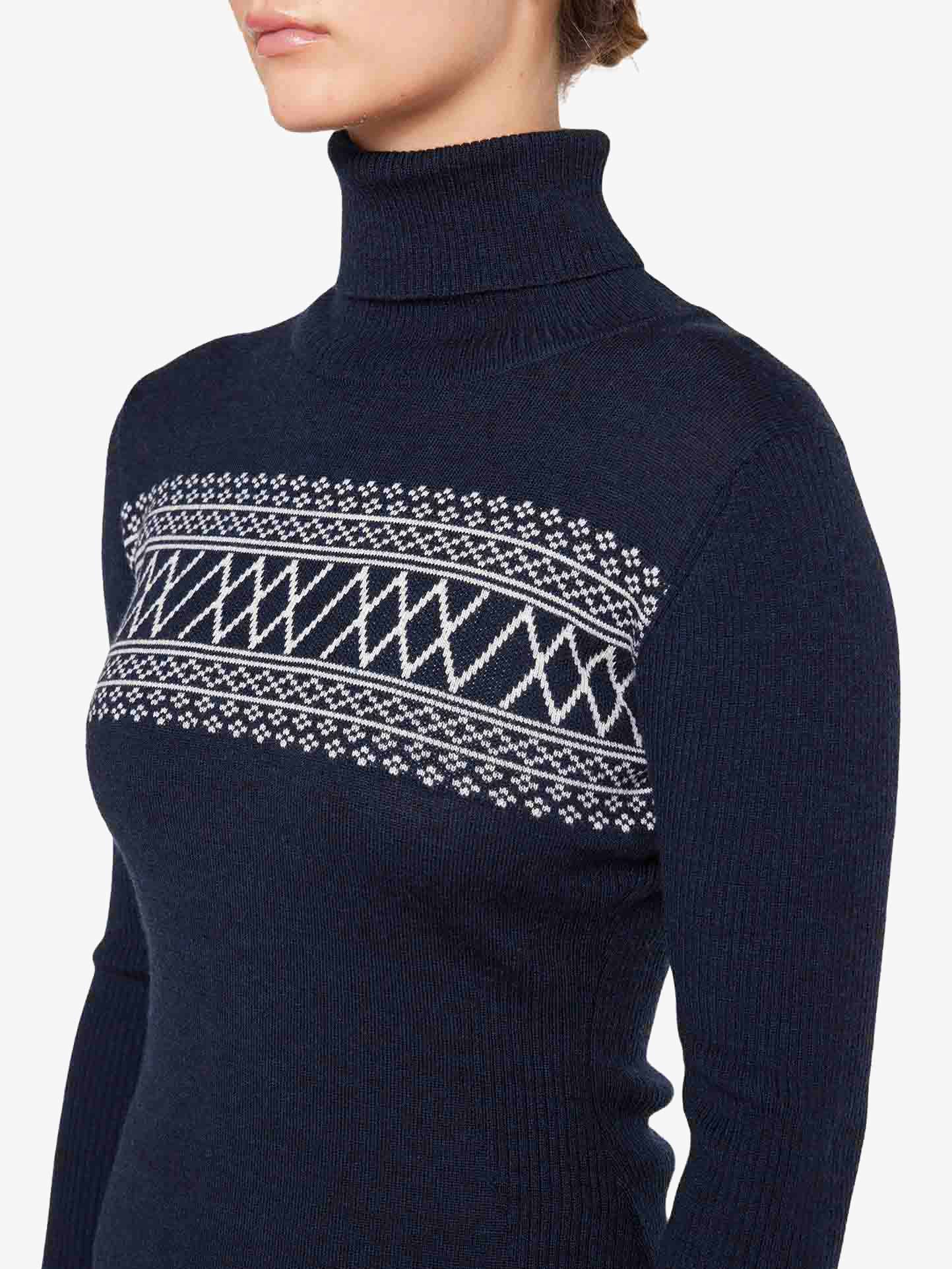 A person wearing the Signature Rollneck Women by We Norwegians, featuring a navy blue design with a striking white geometric pattern across the chest and sleeves. The image captures the upper torso and neck as the person's head is slightly turned to the side, evoking a retro ski outfit aesthetic.