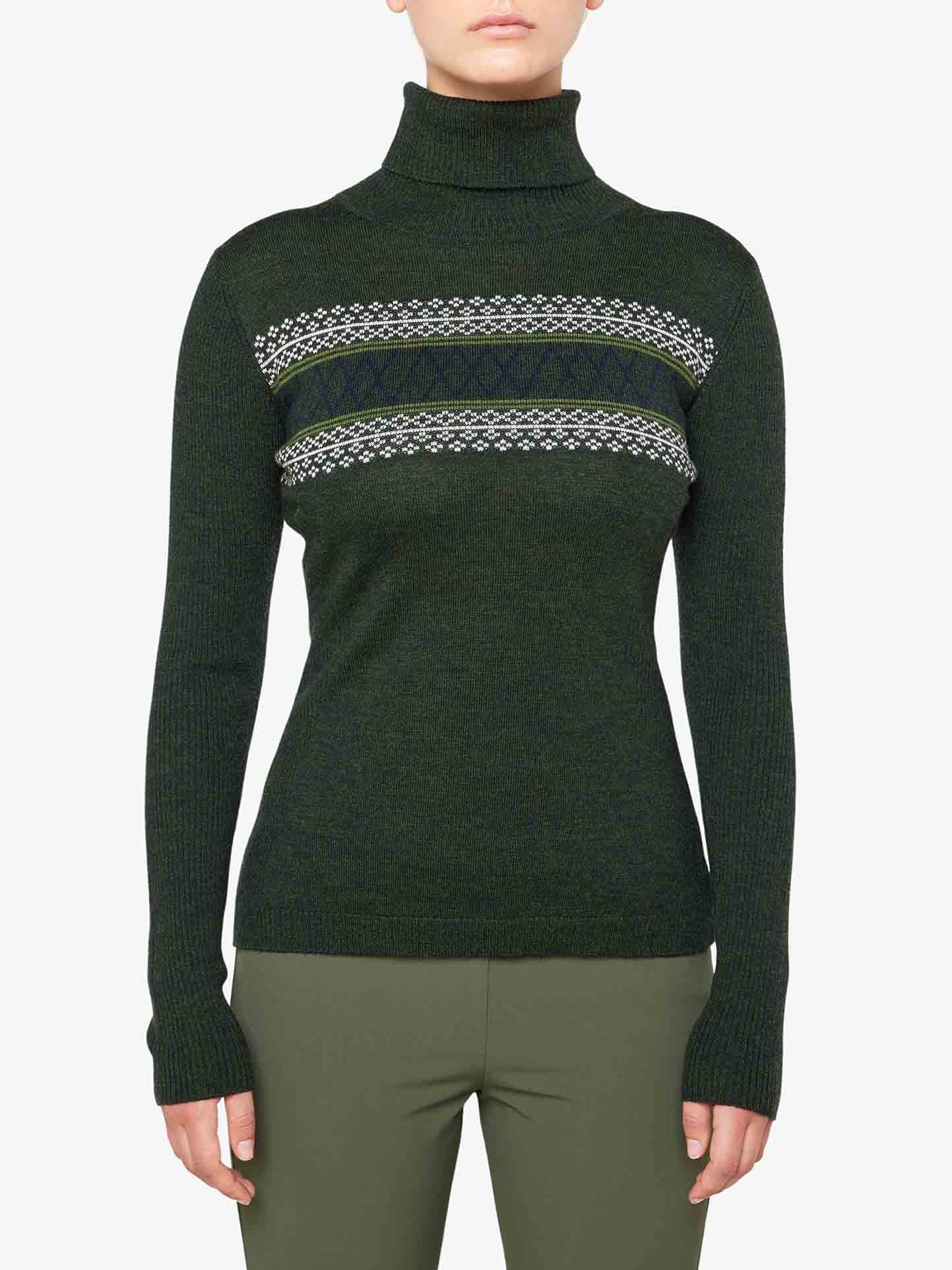 A person is wearing the We Norwegians Signature Rollneck Women sweater in olive green, crafted from fine Merino wool with a patterned knit design across the chest. The sweater boasts long sleeves, and the individual pairs it with matching olive green pants. The plain white background enhances the cozy, retro ski outfit vibes.
