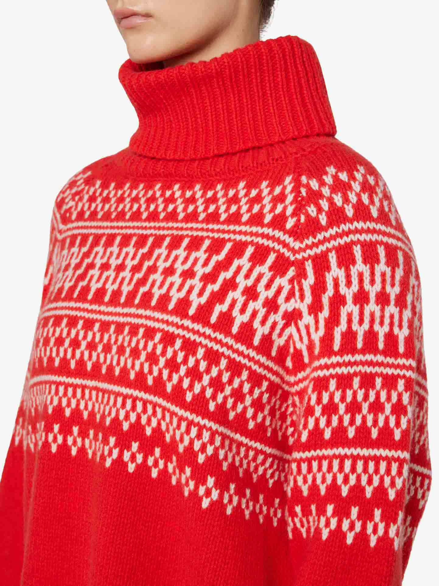 A person wearing the Setesdal Sweater Women by We Norwegians, which is a red knit turtleneck with white patterned detailing. The sweater features intricate, symmetrical designs across the chest and upper arms, showcasing traditional Norwegian knits. The image focuses on the upper torso and a portion of the individual's face.