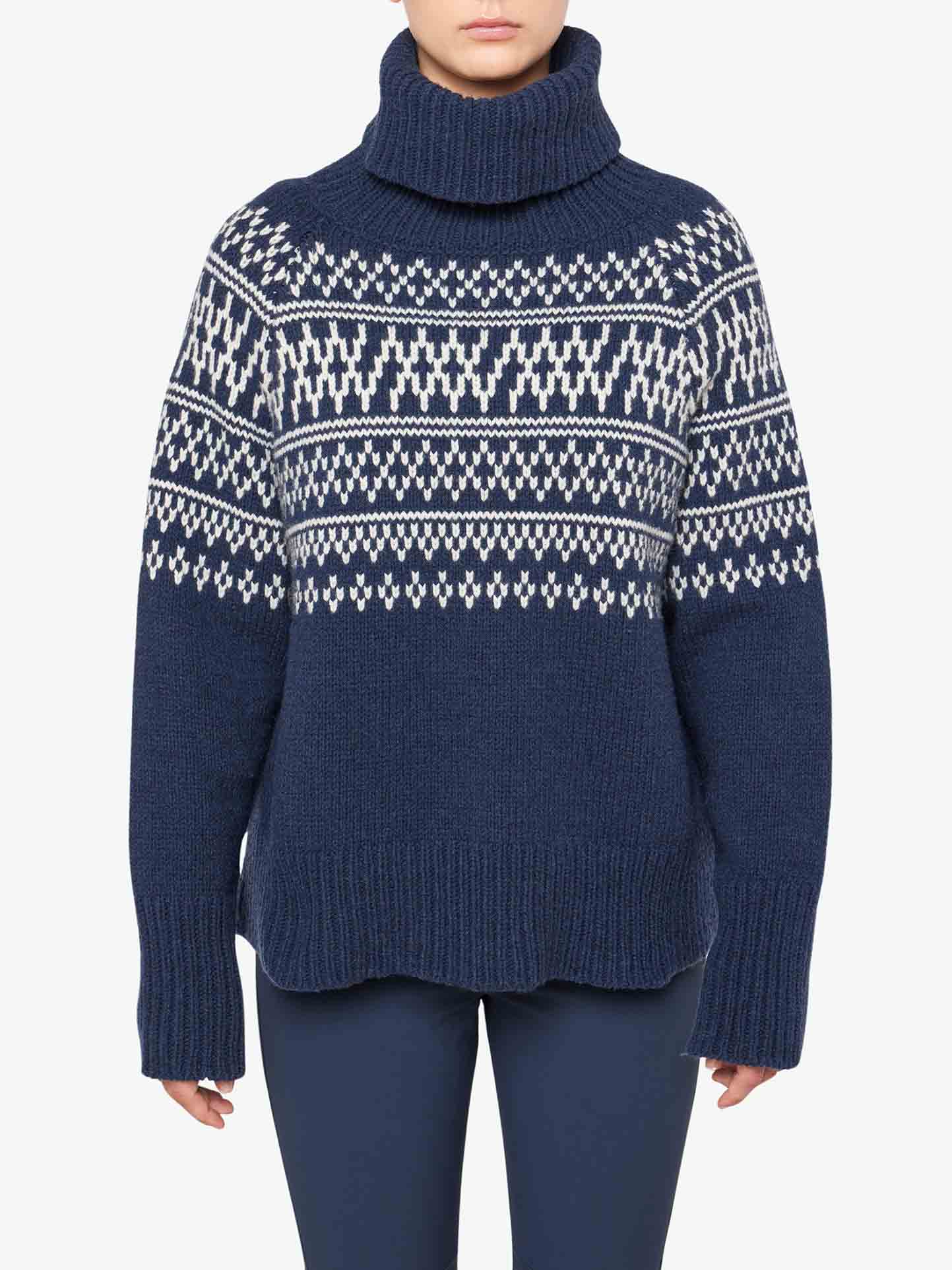 A person wearing a We Norwegians Setesdal Sweater Women in navy blue, featuring a white and blue geometric pattern made from merino/cashmere yarn. The sweater has long sleeves and a loose fit. The individual is also dressed in navy blue pants. The background is plain white.