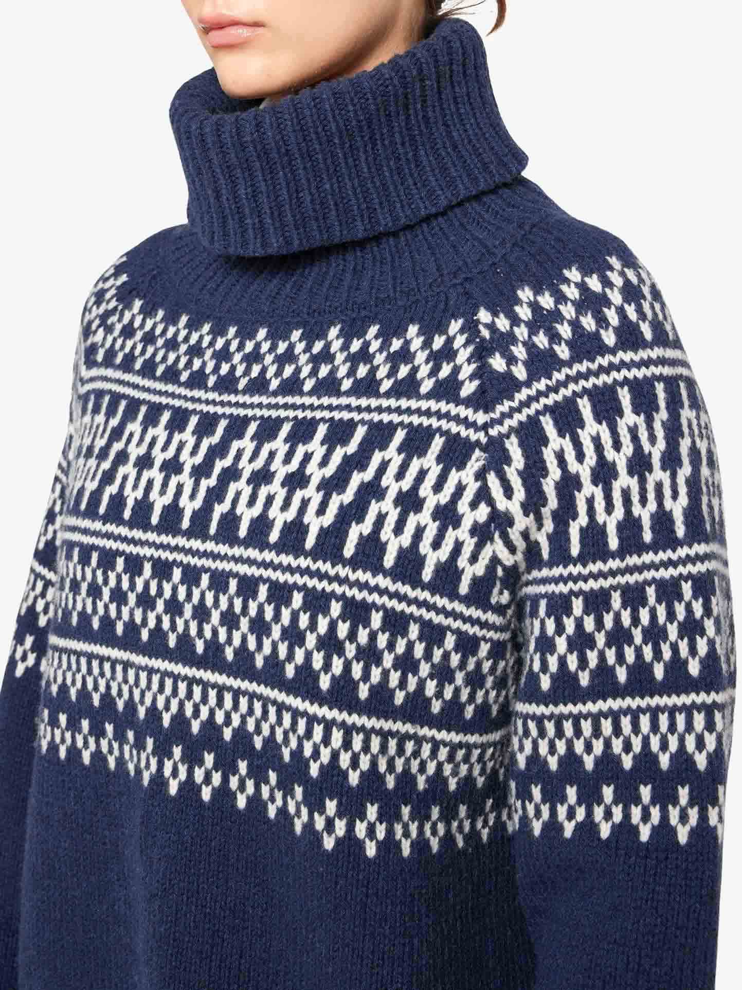 A person is wearing the Setesdal Sweater Women by We Norwegians, a blue and white patterned chunky knit turtleneck sweater made from merino/cashmere yarn. The sweater showcases intricate snowflake-like designs across the chest and shoulders, reminiscent of traditional Norwegian knits. Only the lower half of the person's face is visible.