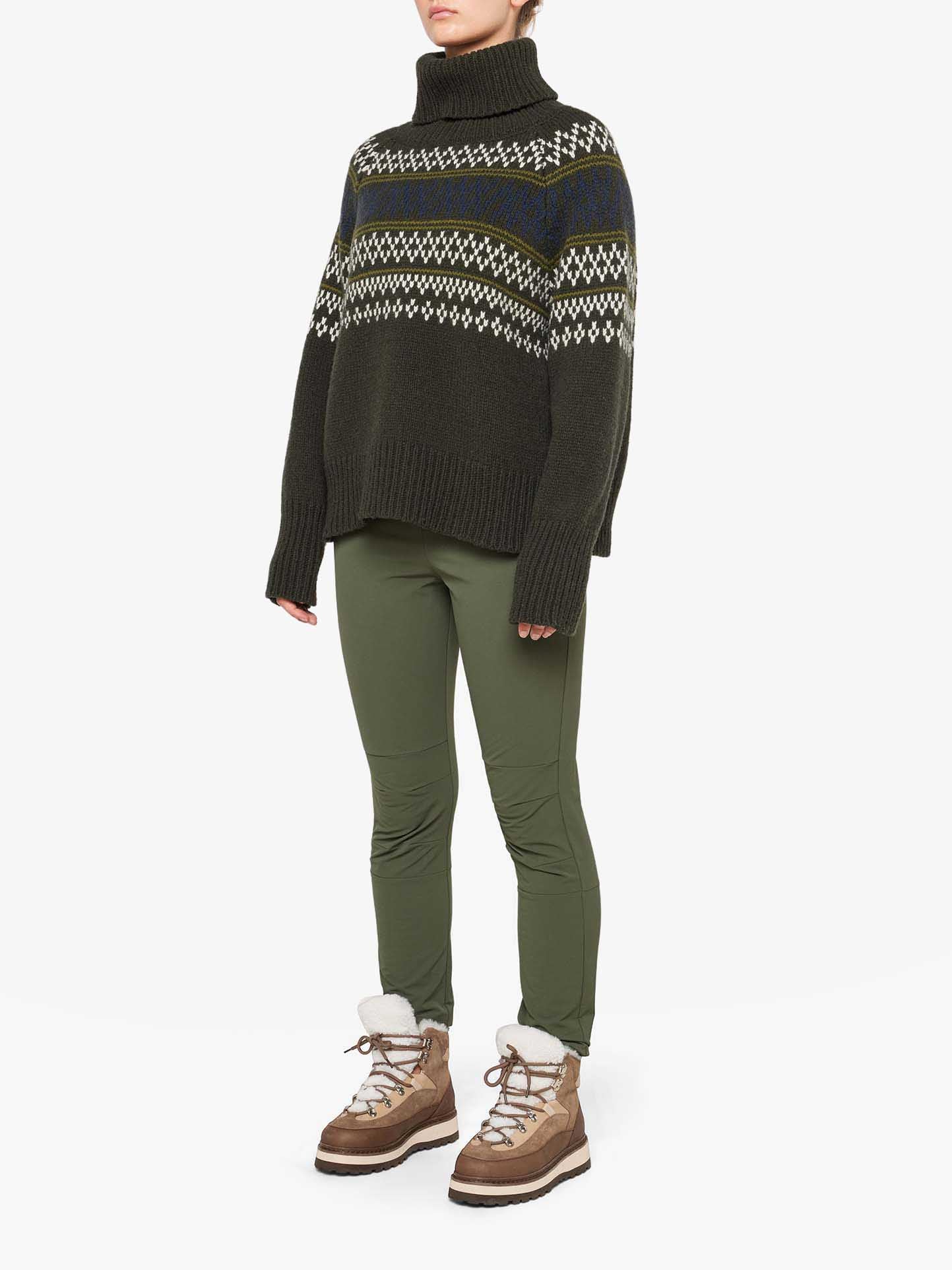 A person is wearing a dark green, patterned Setesdal Sweater Women from We Norwegians paired with olive green pants and light brown winter boots. The boots have white fur trim and are fastened with beige laces. The person is standing against a plain white background.