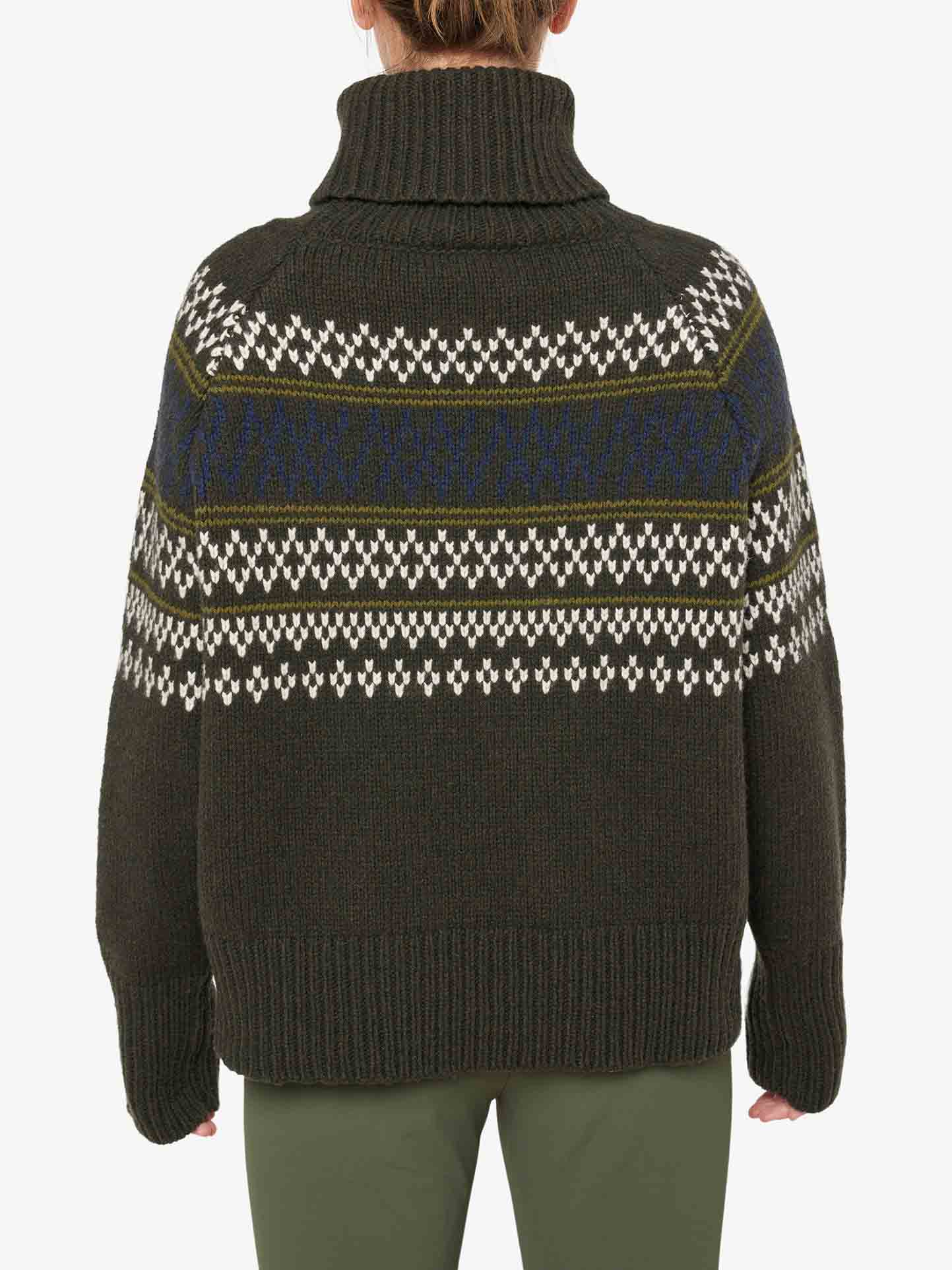 A woman is seen from the back wearing the We Norwegians Setesdal Sweater Women, a luxurious dark green, high turtleneck knitted sweater made from merino/cashmere yarn. The sweater showcases a pattern of white and blue horizontal stripes. She is also wearing green pants.
