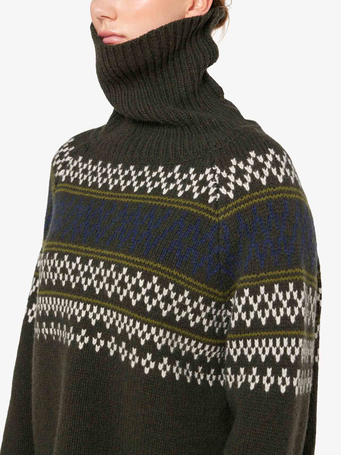 A person is wearing the We Norwegians Setesdal Sweater Women, featuring a high-neck design in dark green with intricate blue, white, and olive green patterns. The cozy sweater boasts a chunky knit texture crafted from soft cashmere yarn. The person's face is partially obscured by the rolled-up collar. The background is plain white.