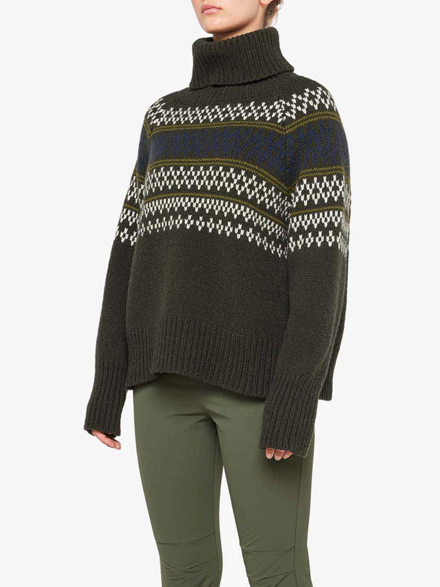 Someone is wearing the We Norwegians Setesdal Sweater Women in dark green, featuring white and blue traditional Norwegian knits across the chest and arms. The sweater has a relaxed fit with long sleeves extending past the wrists. They are also dressed in green trousers, standing against a plain white background.