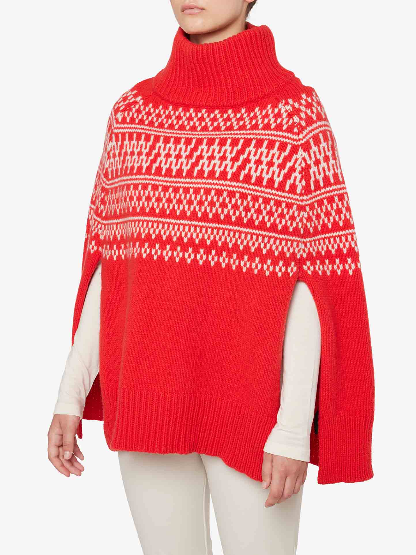 A person wearing a We Norwegians Setesdal Poncho Women with a red and white patterned turtleneck. The poncho, crafted from luxurious merino/cashmere yarn, features a geometric design across the chest and wide-open arm slits. The person is also wearing a white long-sleeve top and white pants underneath.