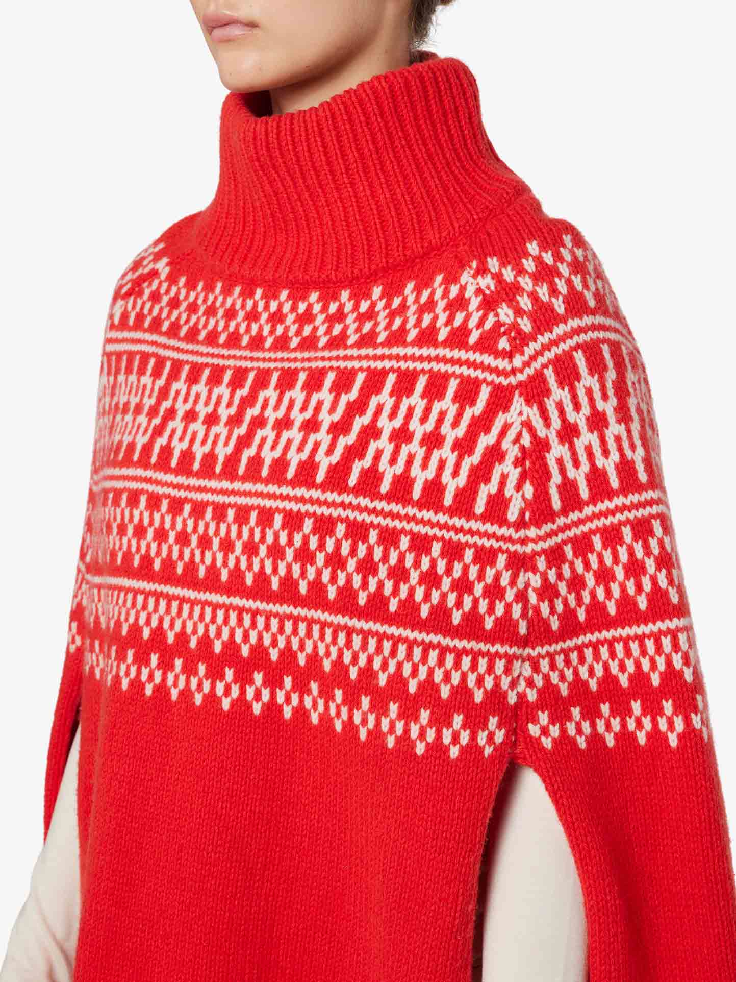 Close-up of a person wearing the We Norwegians Setesdal Poncho Women, made from luxurious merino/cashmere yarn. The red knit poncho features a white, intricate, symmetrical pattern on the yoke and shoulders. It has a high, ribbed turtleneck and the person is visible from the neck to mid-body, angled slightly to the left.