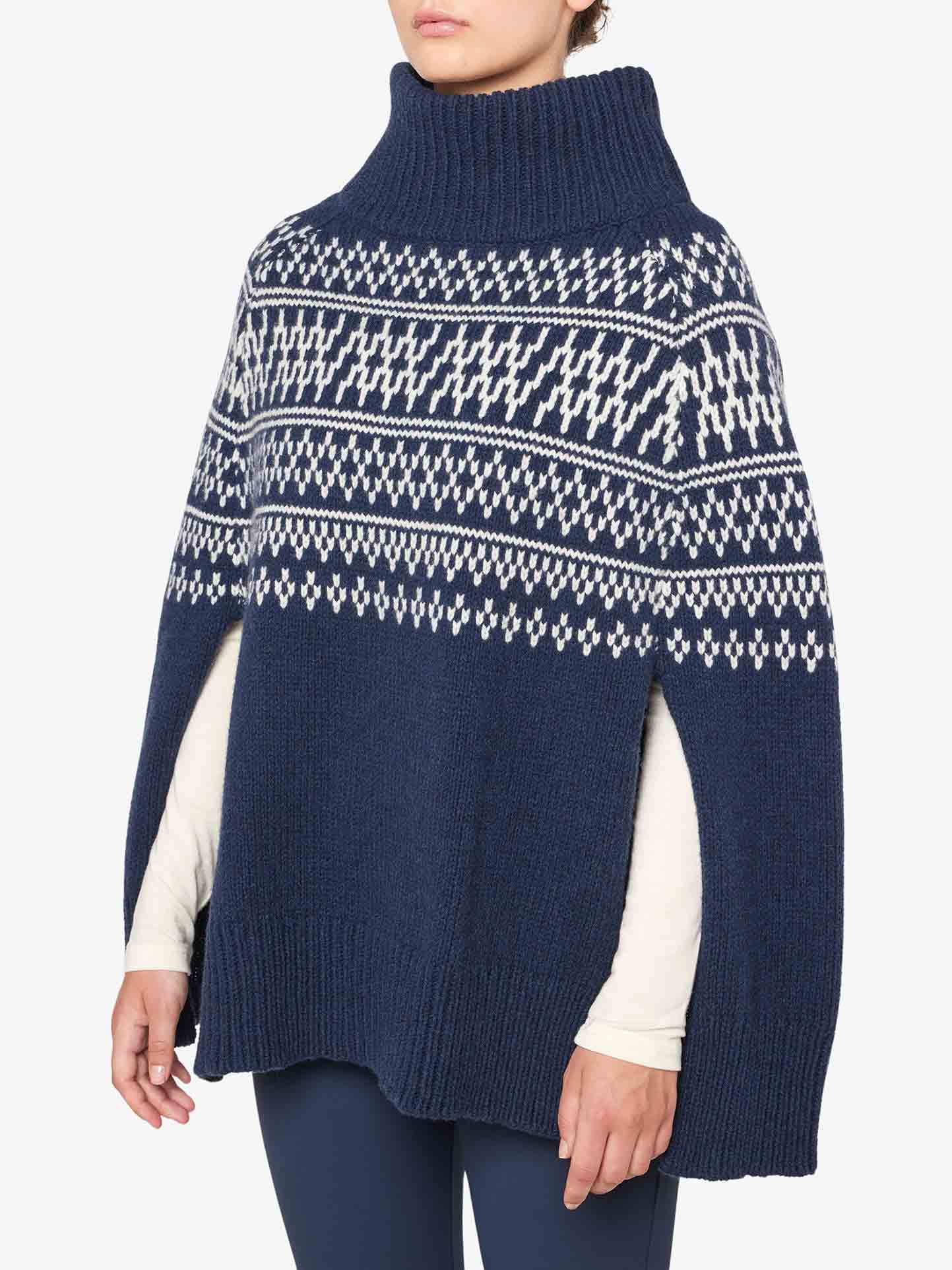 A person is wearing the We Norwegians Setesdal Poncho Women, a blue knitted poncho adorned with traditional Norwegian white and grey patterns. The poncho showcases a high turtleneck and has arm slits, revealing a long-sleeved shirt underneath. The individual is standing against a plain white background.