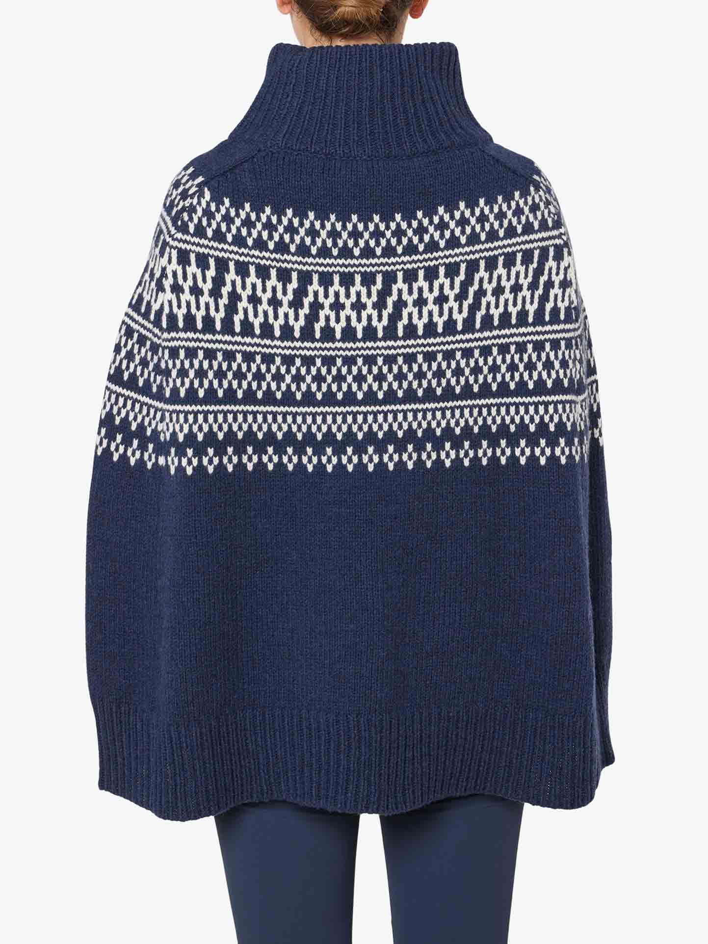 The image shows a person from behind wearing the Setesdal Poncho Women by We Norwegians, a dark blue, high-neck poncho with a white Nordic pattern across the upper back, reminiscent of traditional Norwegian knits. The poncho appears oversized, covering primarily the back and part of the arms, and the person is also wearing dark blue pants.