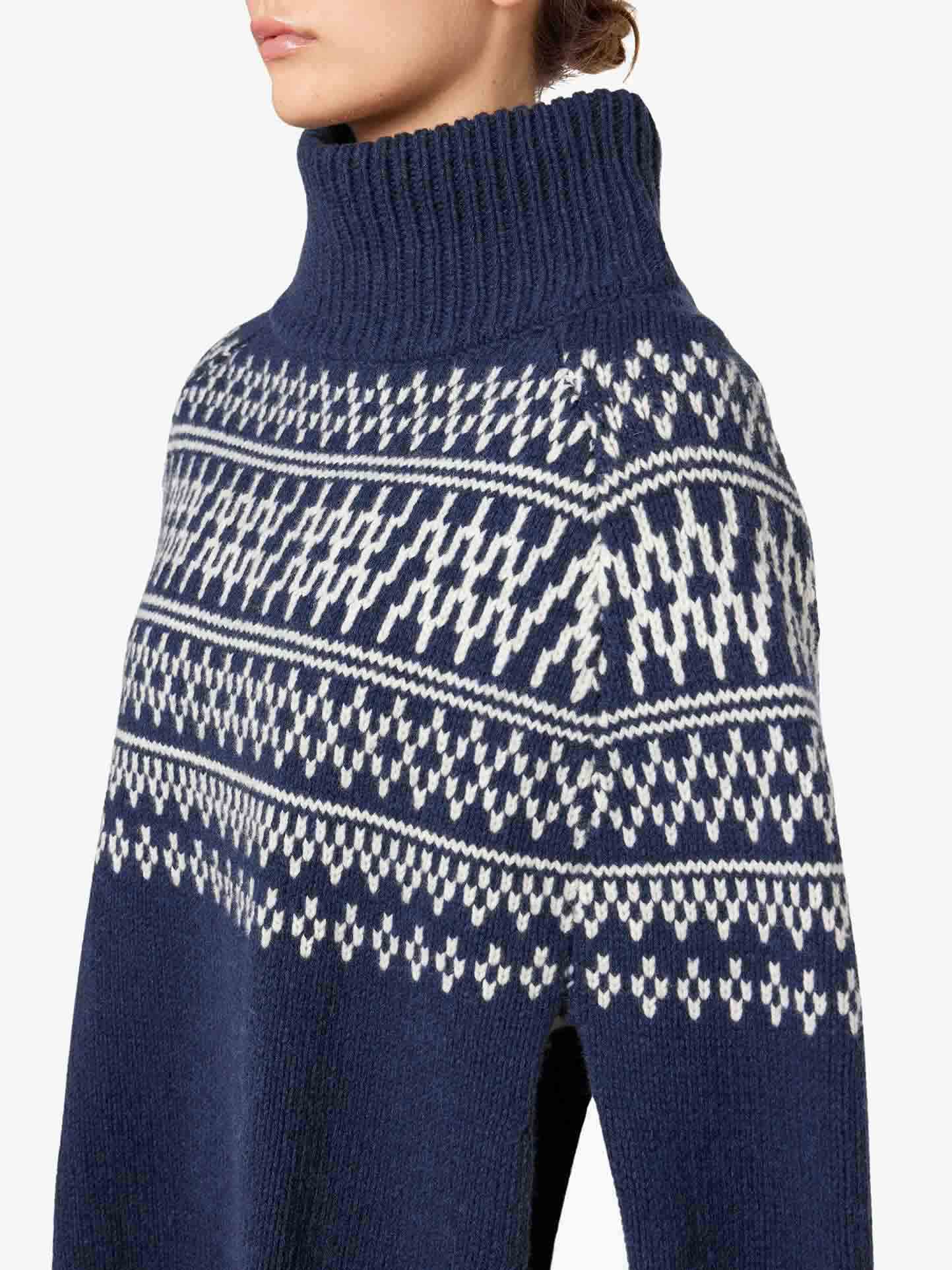 A person wearing the Setesdal Poncho Women by We Norwegians, a dark blue garment adorned with white patterned knit details. Inspired by traditional Norwegian knits, this poncho showcases a festive and intricate design across the chest and shoulders. The individual is shown from the neck down, set against a white background.