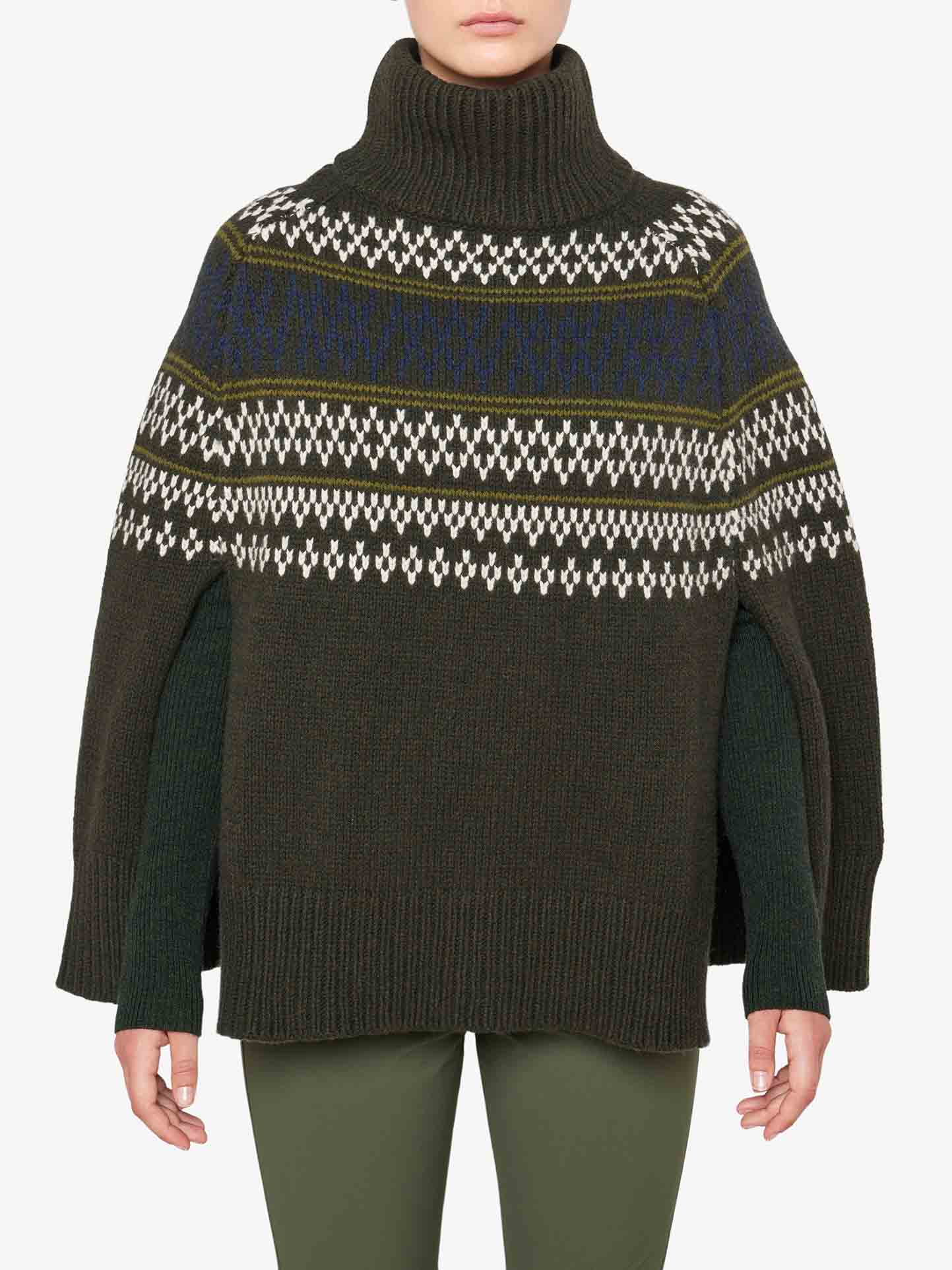 A person wearing the Setesdal Poncho Women by We Norwegians, an olive green turtleneck poncho sweater adorned with a Fair Isle design in cream and blue across the chest and upper arms. Crafted from luxurious merino/cashmere yarn, the sweater features an oversized fit with side slits that reveal long sleeves underneath. The person is also wearing green pants.