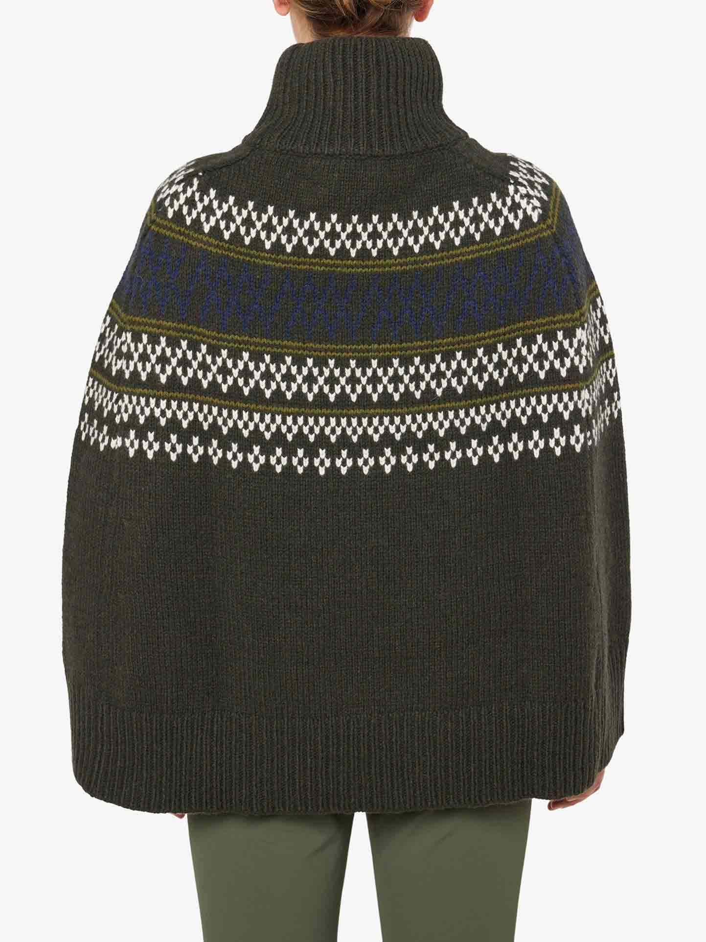 Back view of a person wearing the Setesdal Poncho Women by We Norwegians, featuring a high neck and dark green knit fabric with a traditional Norwegian patterned design in white and blue across the shoulders. The poncho drapes loosely over the torso with a ribbed hem, complemented by matching green pants.