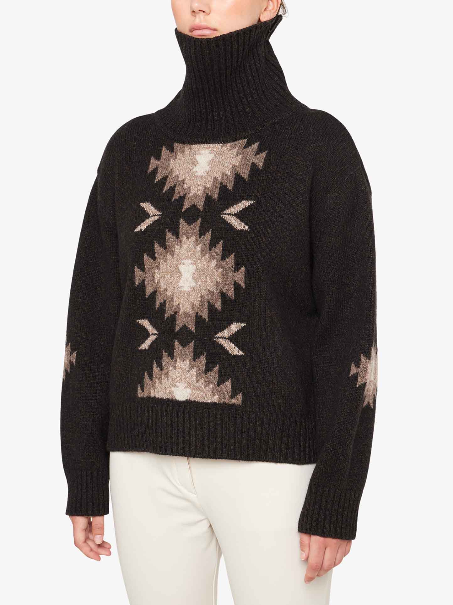 A person is wearing the Hovden Sweater Women by We Norwegians, a dark, oversized turtleneck adorned with geometric patterns in light brown and beige, evoking Norwegian Sámi cultural designs. The cozy roll neck features a ribbed collar, cuffs, and hem. They are also dressed in light-colored pants. The person's head is partially cropped from the image.