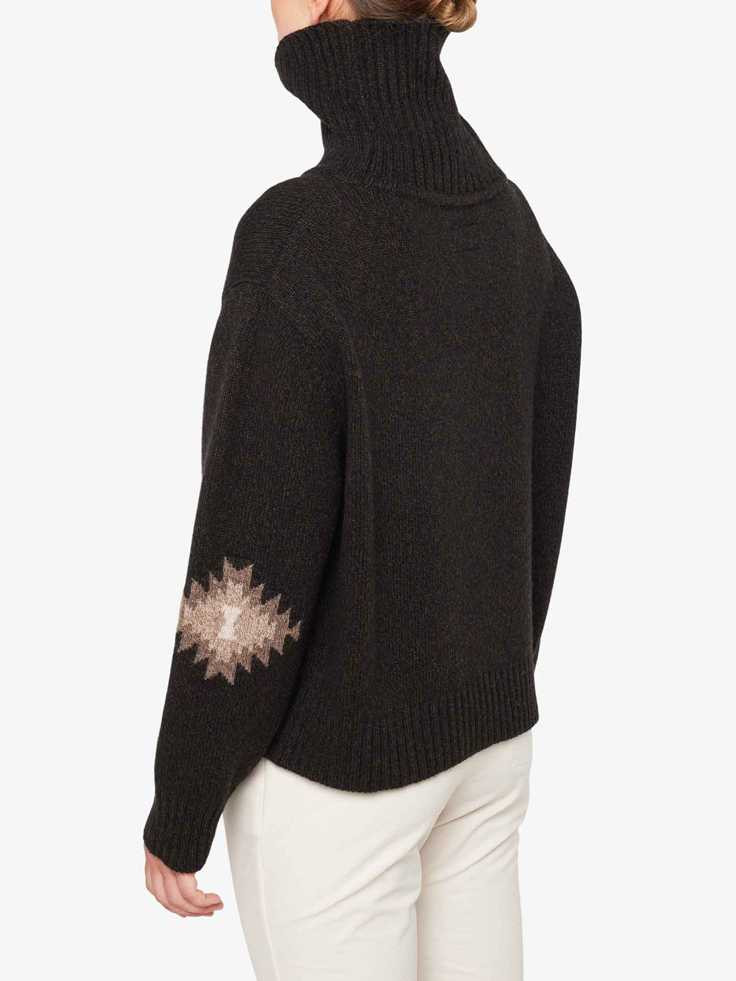 A person wearing the Hovden Sweater Women by We Norwegians, featuring a cozy roll neck and a beige geometric pattern on one sleeve, is seen from the back. They have light-colored pants and their hair is tied up. The background is plain white.