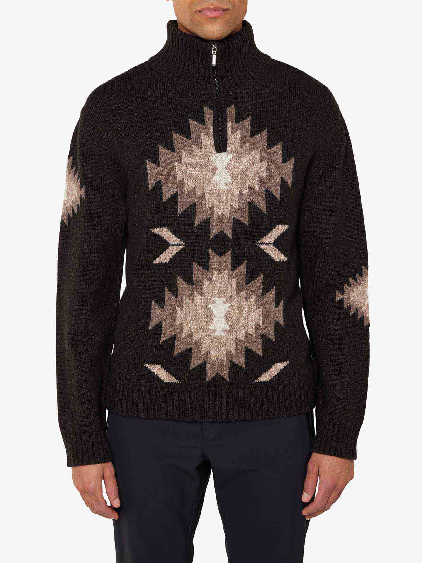 A person wearing the Hovden Sweater Men by We Norwegians, a dark wool sweater with a high collar and a half-zipper, perfect for fall weather. The sweater features a large central geometric pattern in beige and light brown tones with smaller patterns on either side, inspired by Norwegian Sámi culture. The person is also wearing dark trousers.