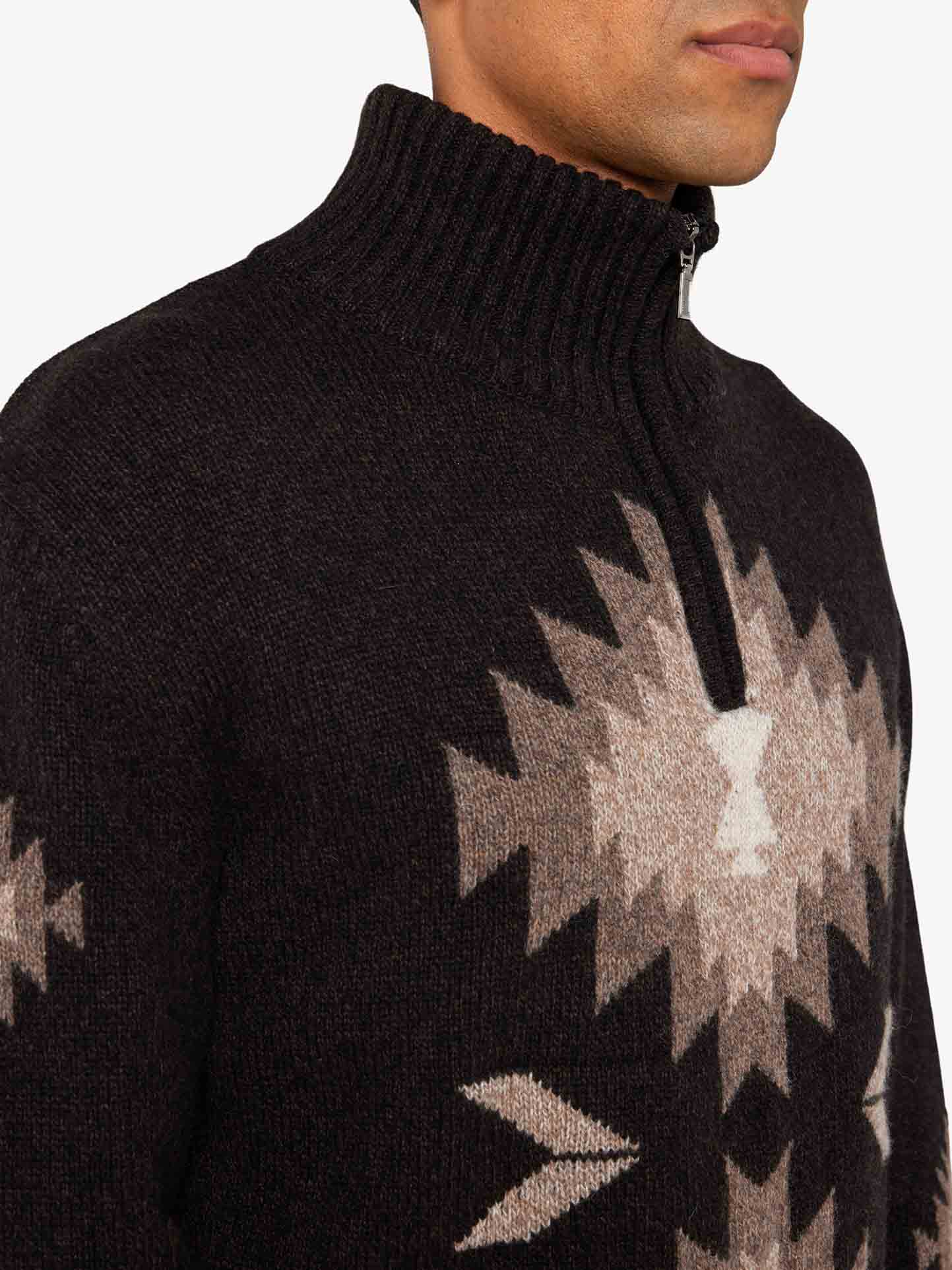 A close-up image of a person wearing the "Hovden Sweater Men" by We Norwegians, a dark, thick, knitted wool sweater with a high ribbed collar and partial zip. The sweater showcases a geometric, diamond-like pattern in lighter tones on the chest and shoulder areas. Perfect for Fall weather, this piece evokes the timeless charm of Norwegian Sámi culture. The background is plain white.