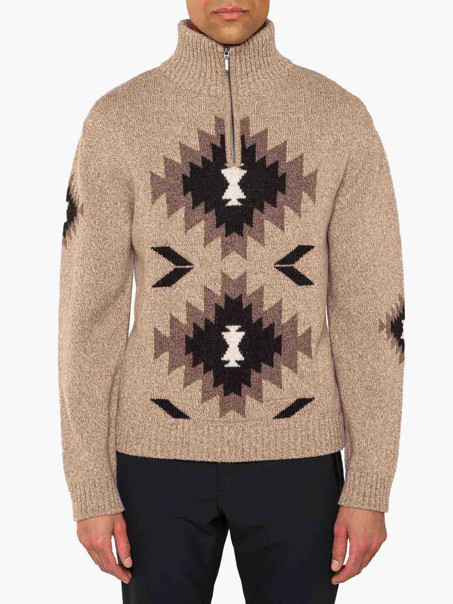 A person wearing the Hovden Sweater Men by We Norwegians, a beige, long-sleeved knitted wool sweater with a high collar and quarter zip. The sweater features a black and brown geometric pattern inspired by Norwegian Sámi culture on the front and sleeves. The person is standing with their arms down, wearing dark pants—an ideal outfit for Fall weather.