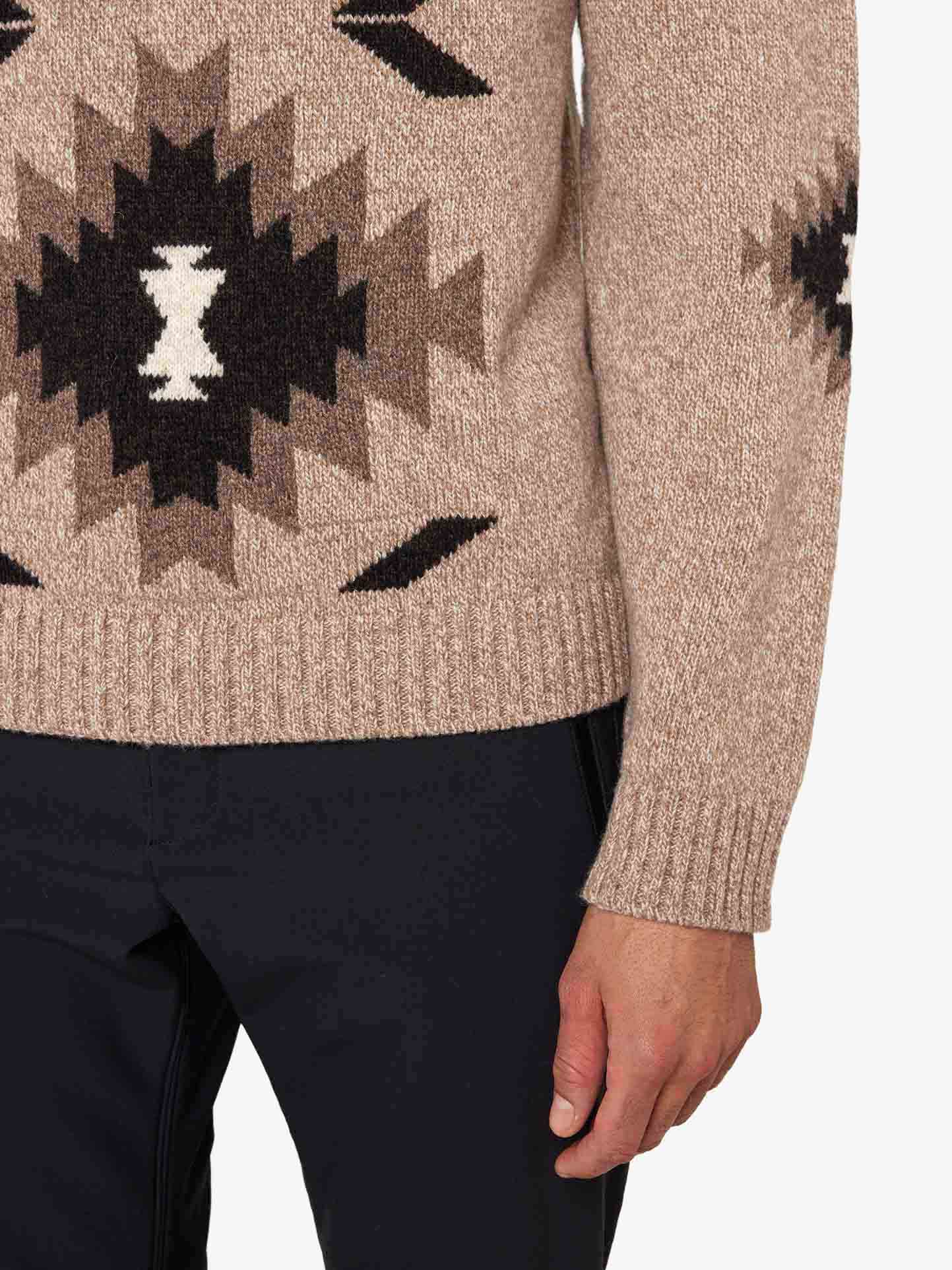 A close-up of a person wearing the Hovden Sweater Men by We Norwegians, featuring a beige knitted wool design with black, white, and brown geometric patterns inspired by Norwegian Sámi culture on the side and sleeve. The person is also wearing black pants, with their right hand visible—perfect for fall weather.