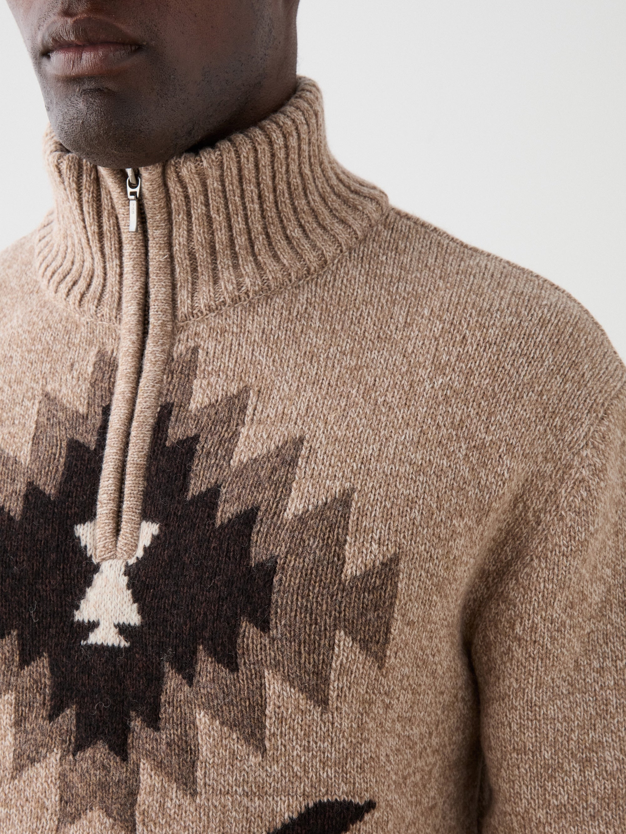 A close-up of a person wearing the We Norwegians Hovden Sweater Men, a beige quarter-zip with a high ribbed collar and geometric black and brown chest pattern. The person's face is partially visible.