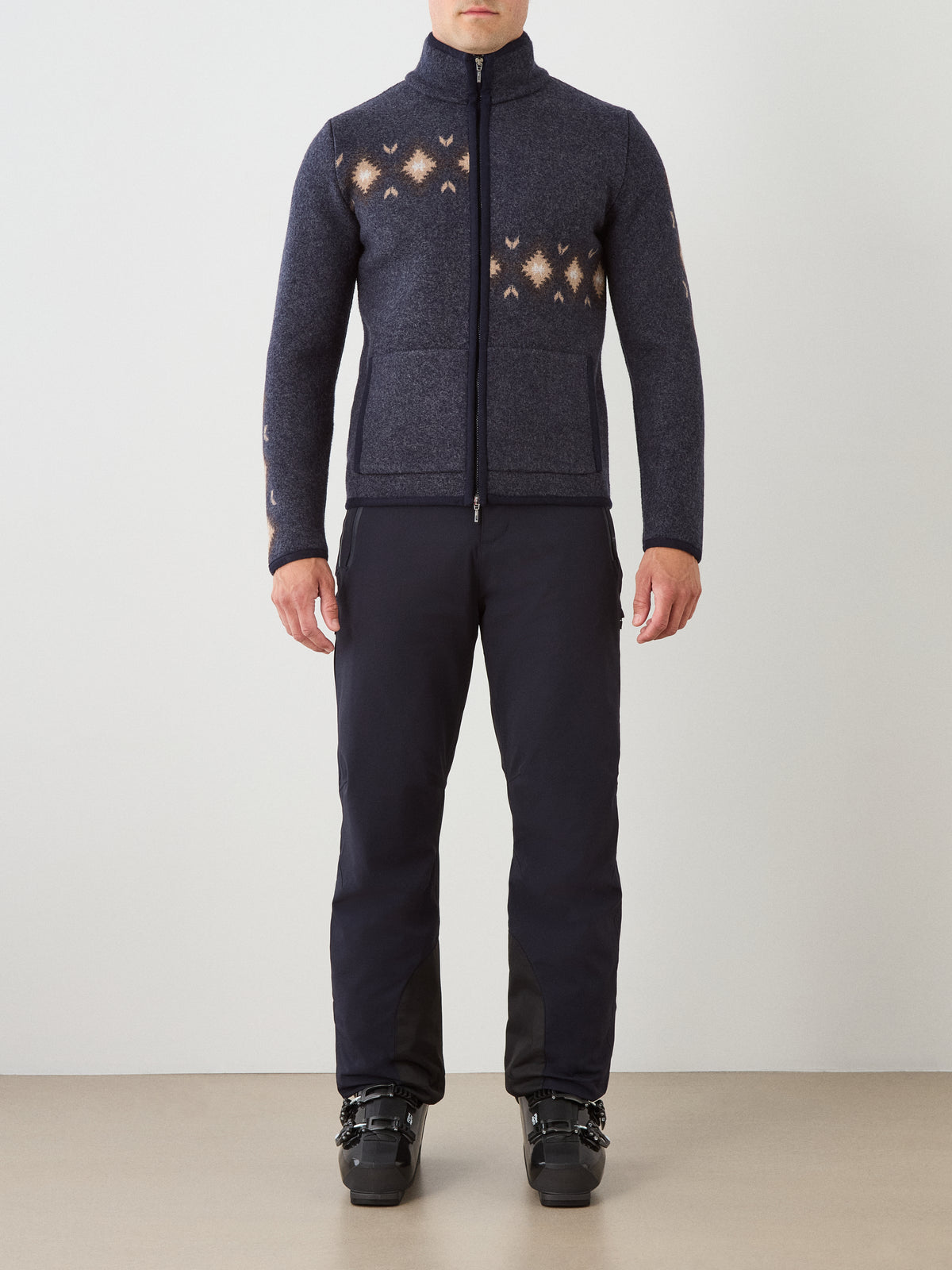 A person wearing the We Norwegians Hovden Jacket Men—a luxury midweight layer in dark boiled merino wool with subtle Norwegian Sámi patterns and a zip-up front—stands facing forward against a light background, paired with black pants and black ski boots.